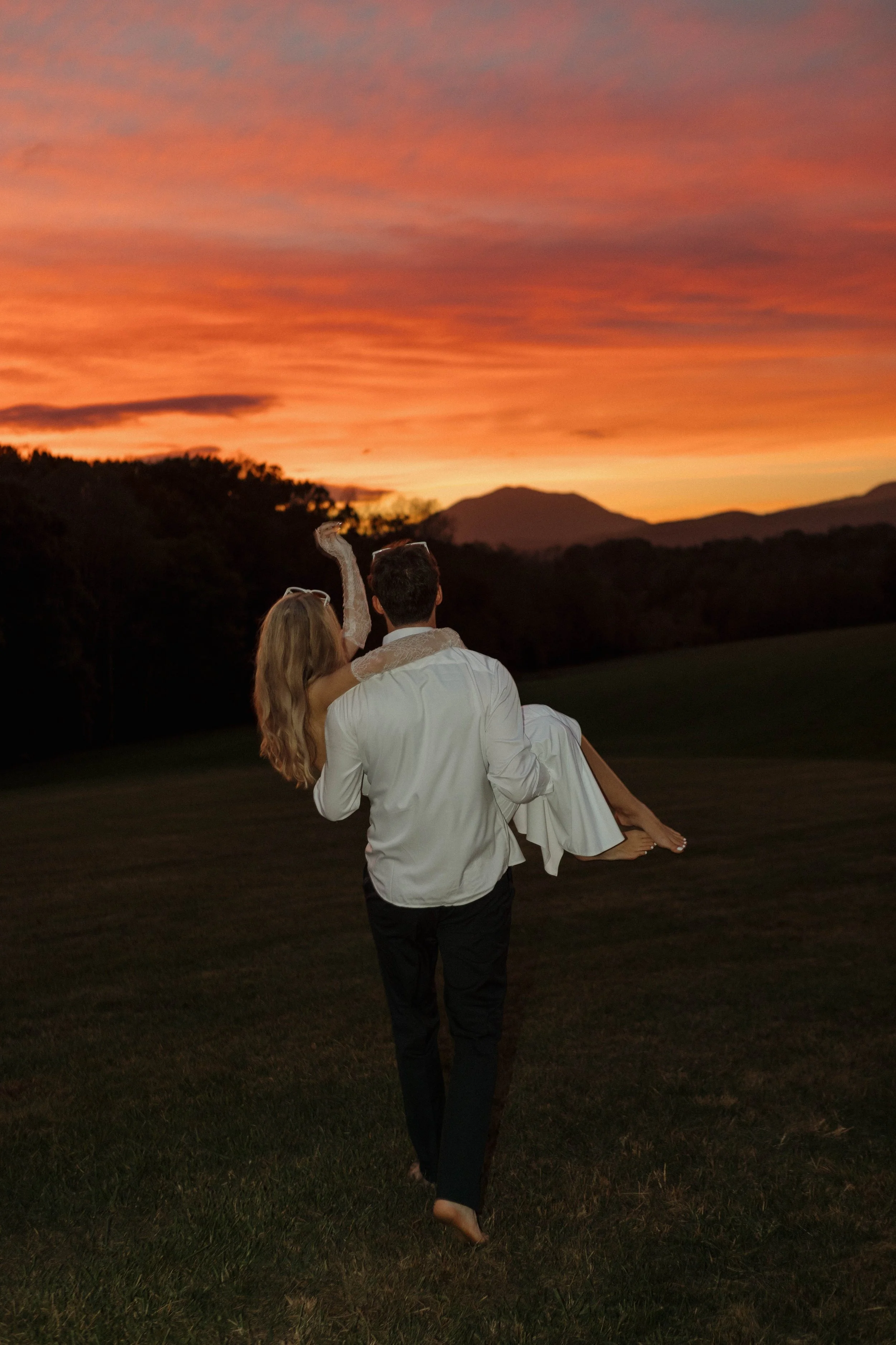 A couple, with the man carrying the woman, walking on grass during a colorful sunset with mountains in the background.