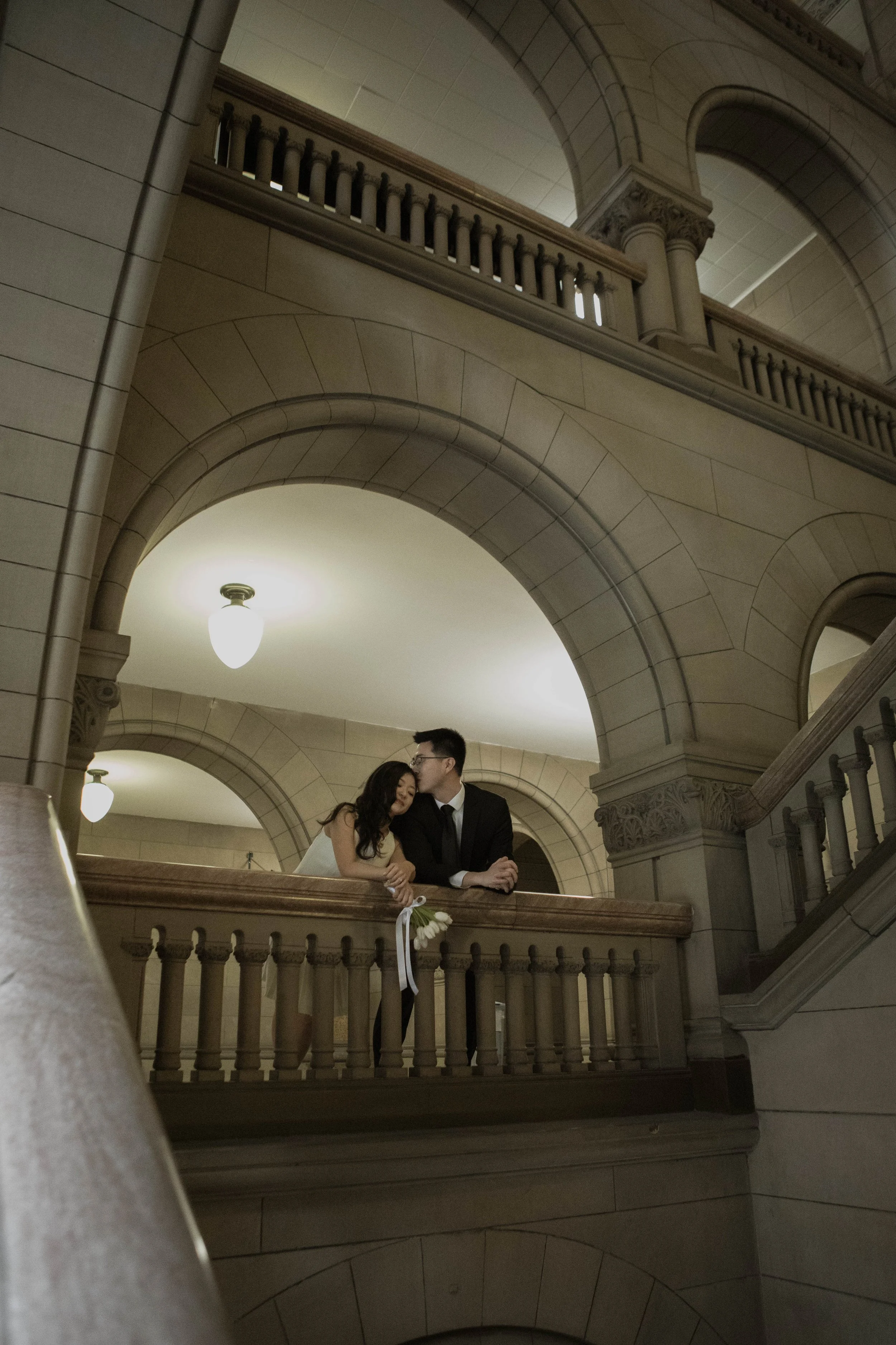 A couple, dressed in formal attire, stands close together on a balcony inside a building with ornate stone architecture, arches, and warm lighting.