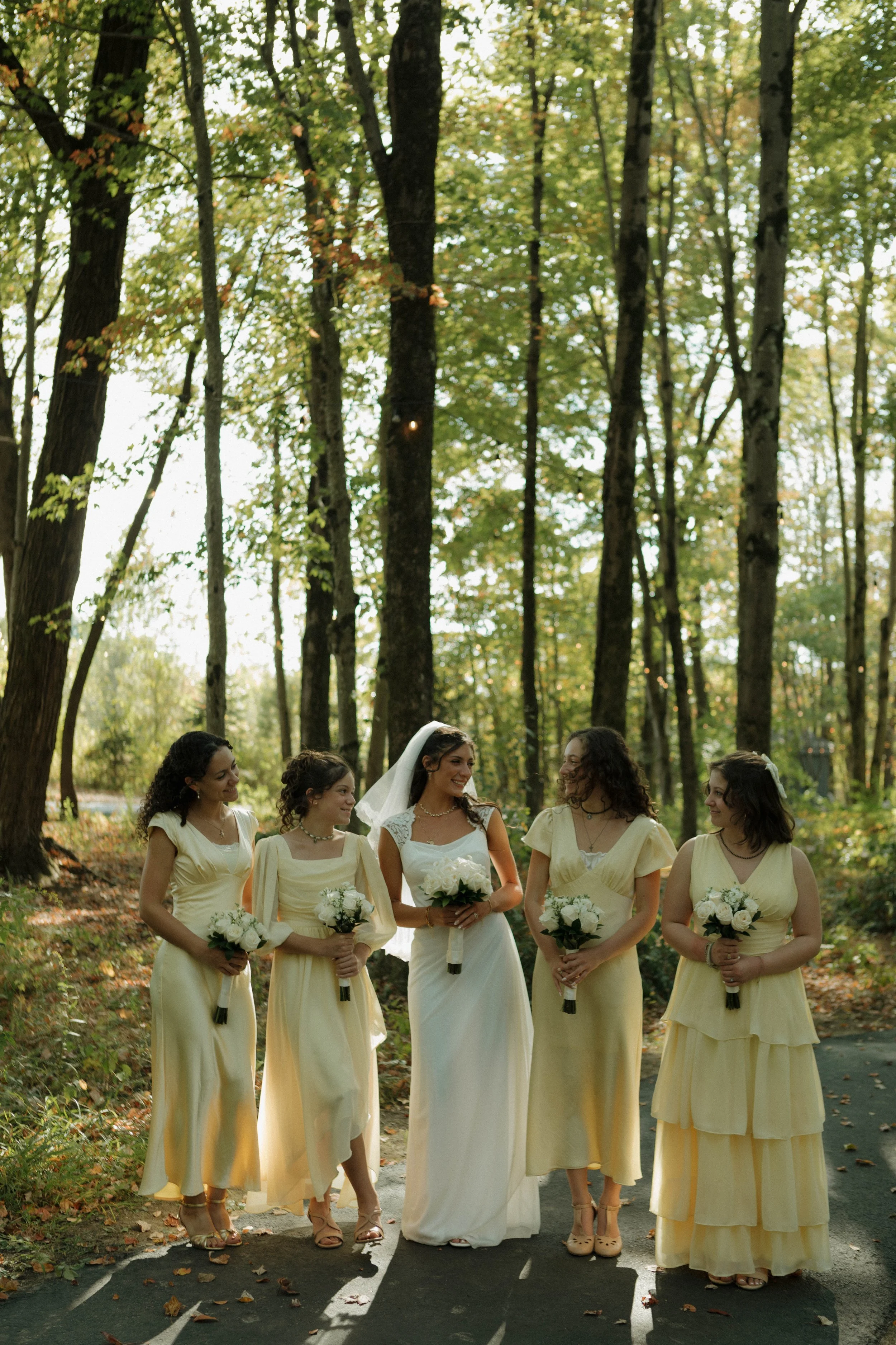 A bride and five bridesmaids in yellow and white dresses walking in a wooded area, holding white bouquets.