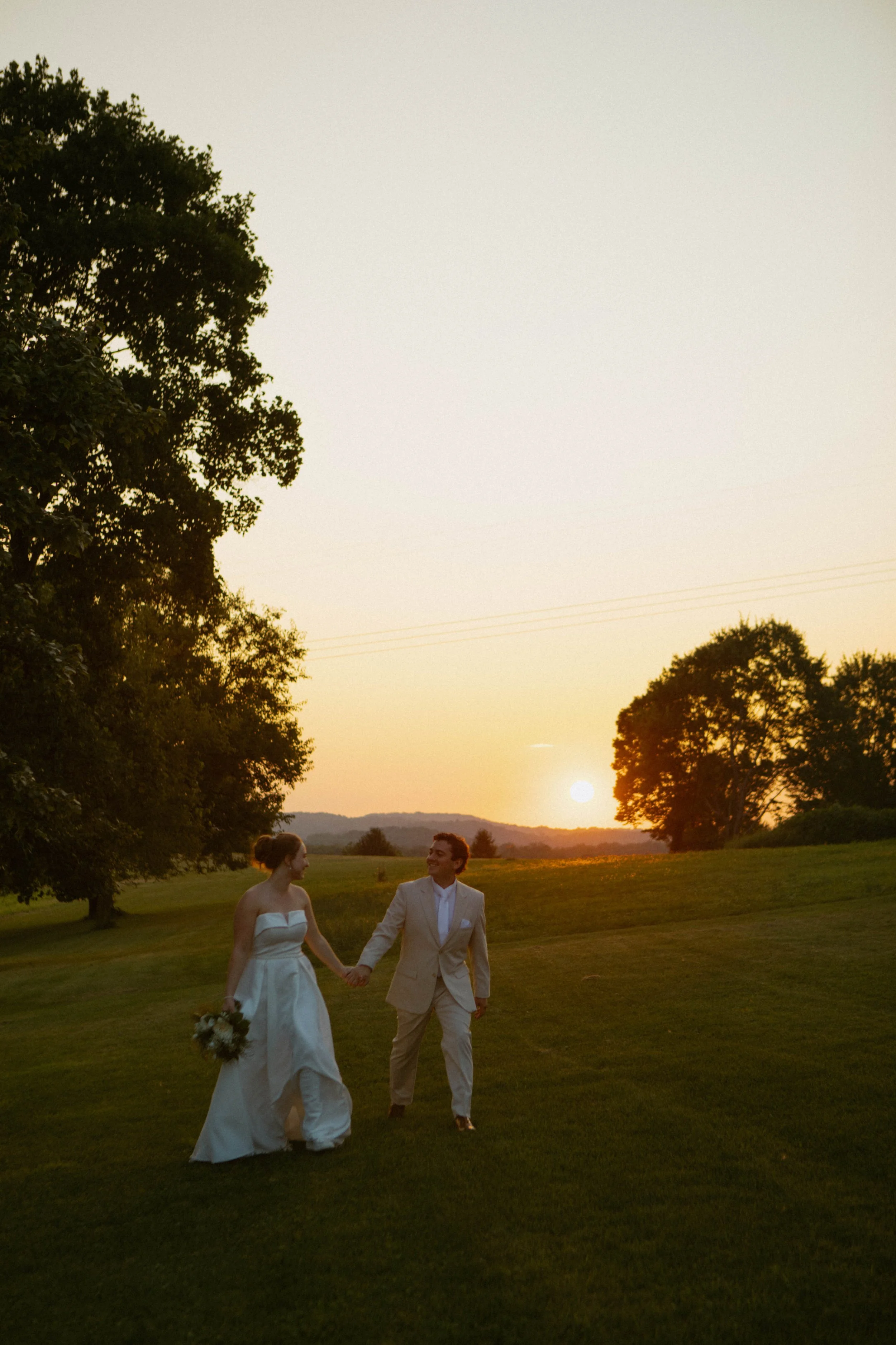 A couple dressed in wedding attire holding hands and walking on a grassy field during sunset, with trees and hills in the background.