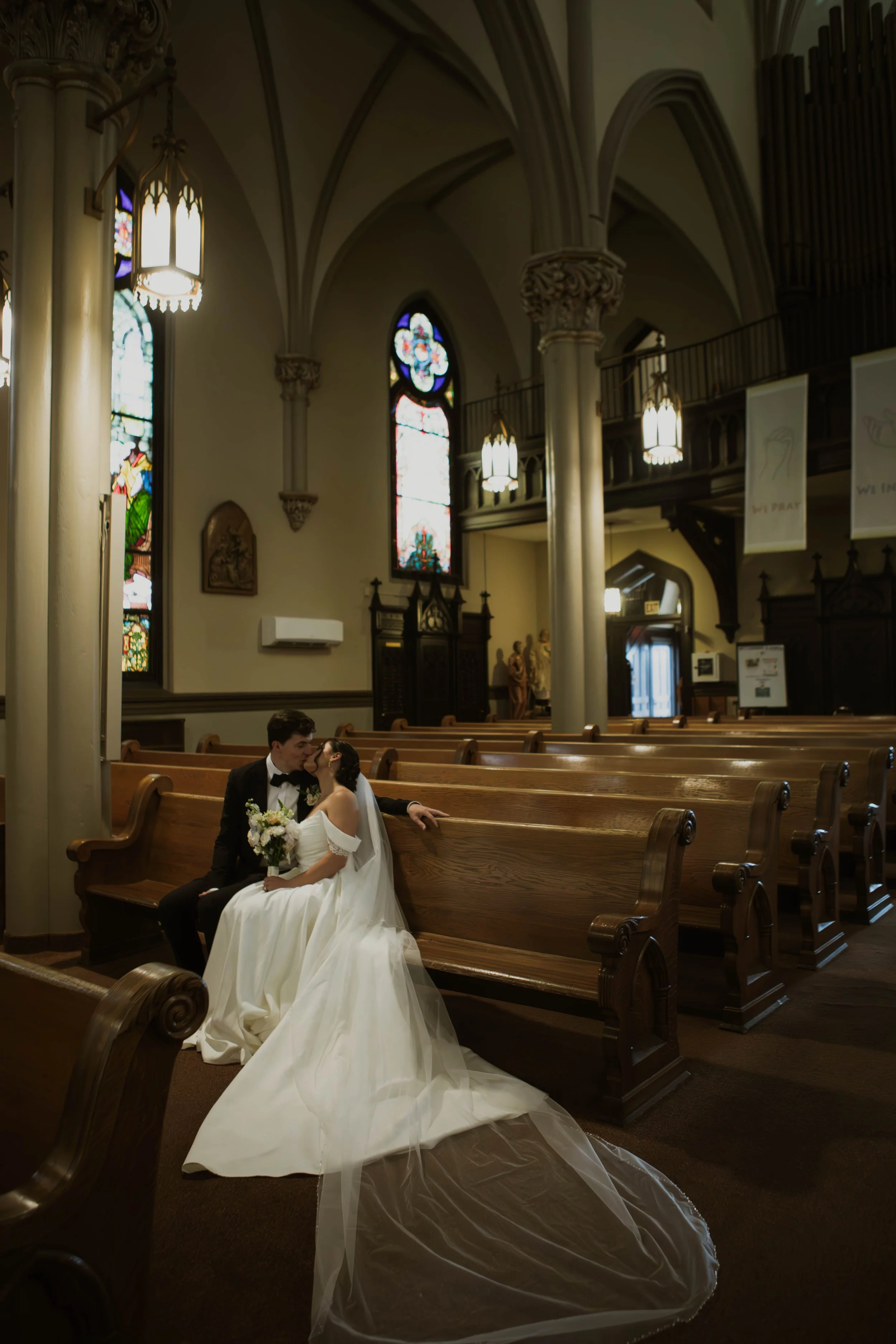 A bride and groom sitting in a church, sharing an intimate moment, surrounded by stained glass windows and wooden pews.