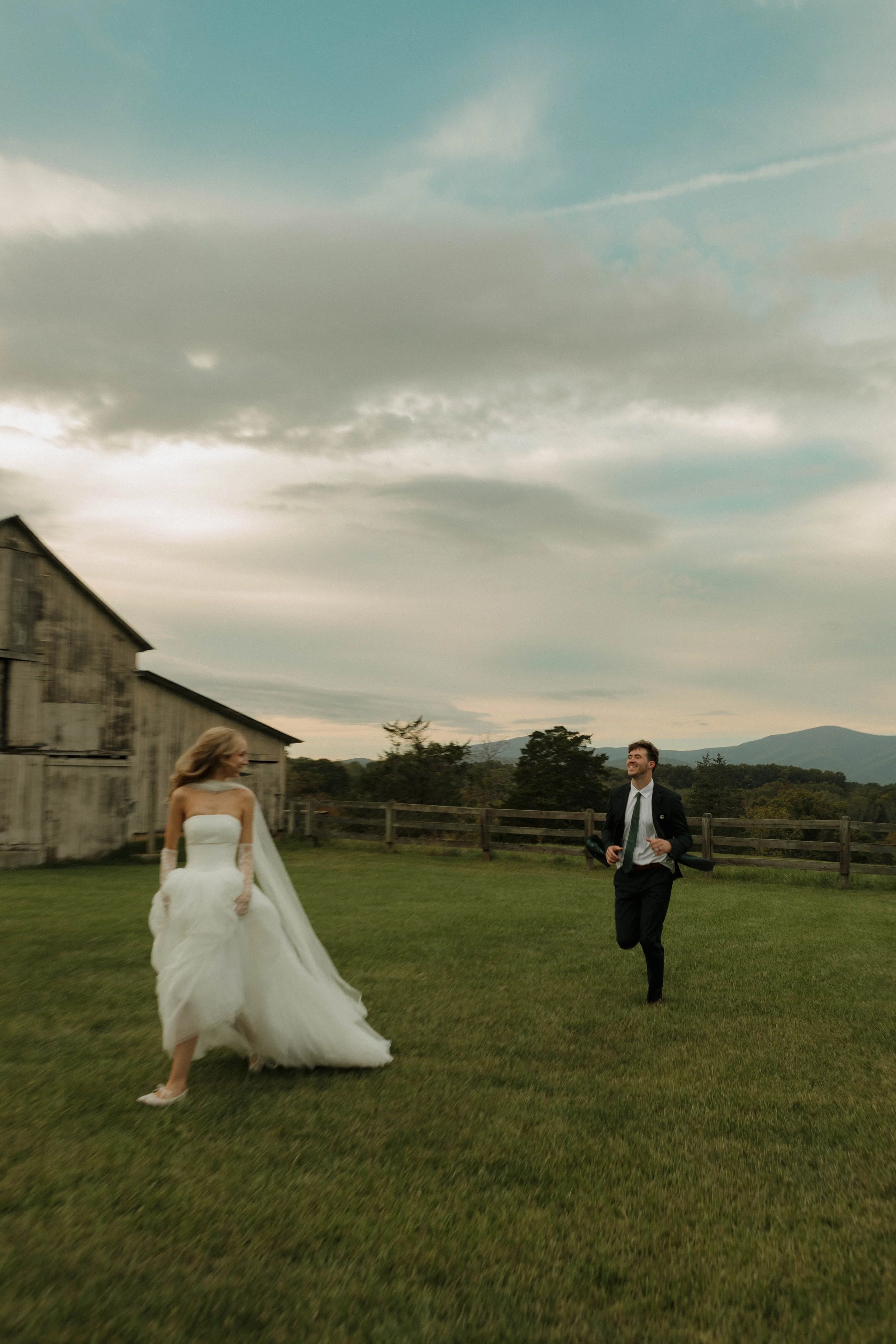 A bride in a white wedding gown and a groom in a black suit running on a green grassy field near a rustic wooden barn, with a cloudy sky and distant hills in the background.
