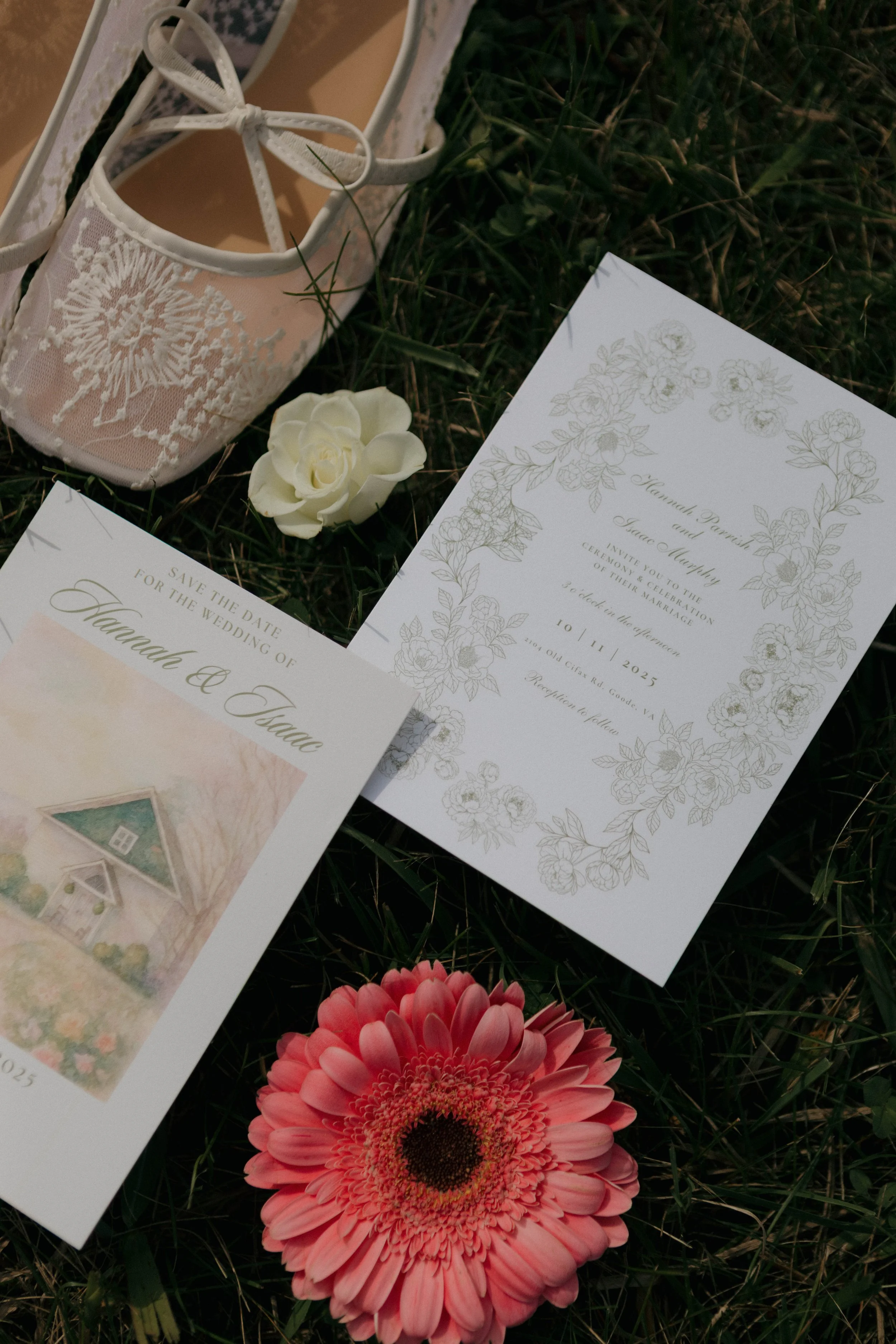 Wedding invitation suite with floral design, a single white flower, a pair of lace shoes, and a pink gerbera daisy laying on grass.