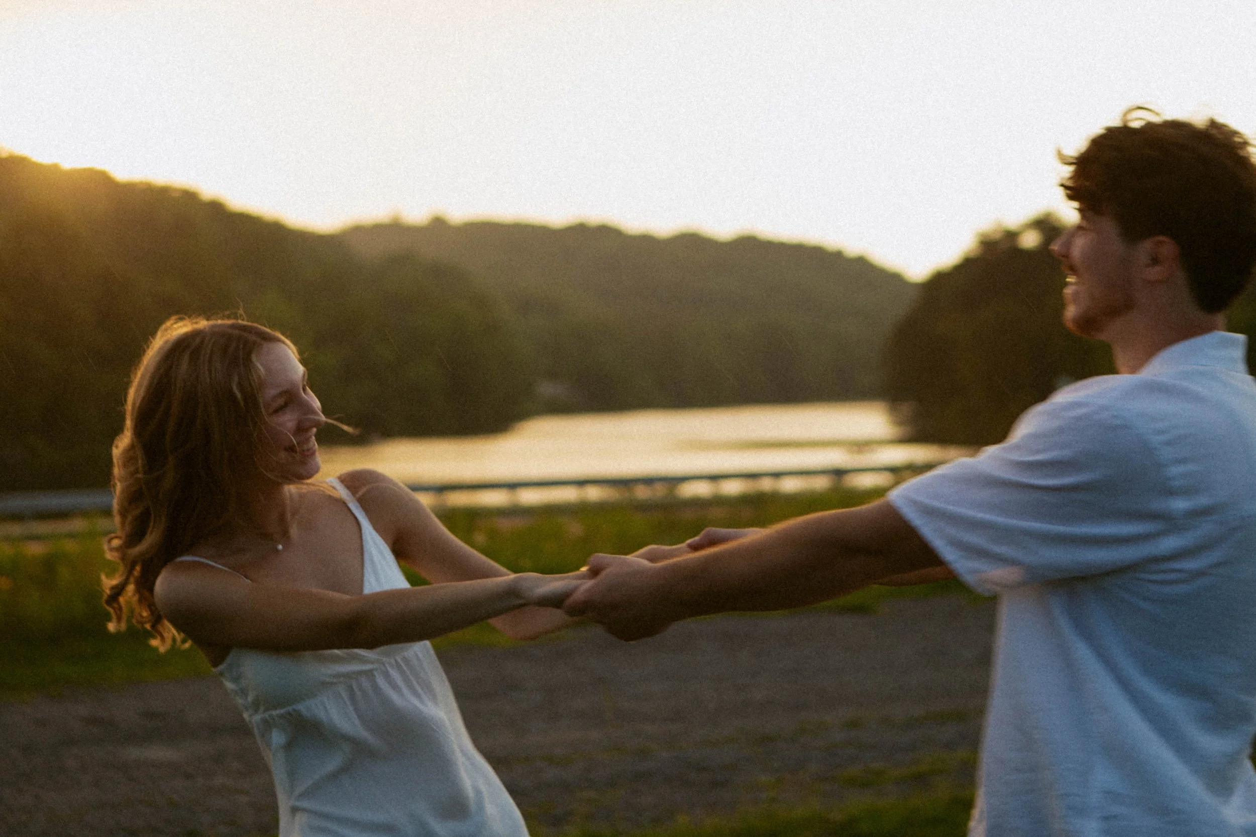 A young couple holding hands and enjoying a moment together outdoors at sunset, near a body of water with hills in the background.