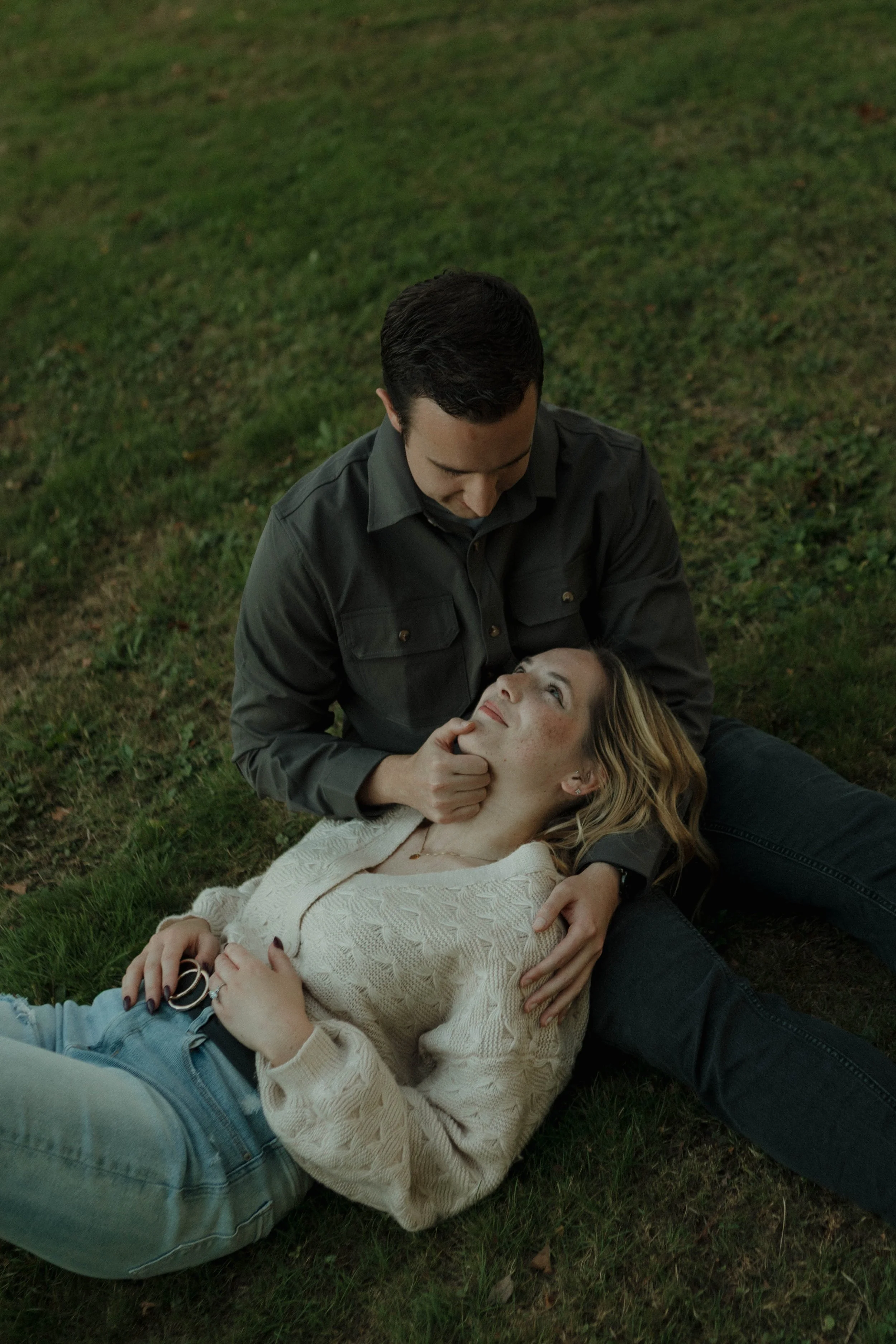 A man and woman are sitting on grass, with the woman lying down and the man leaning over her, looking at her with affection.