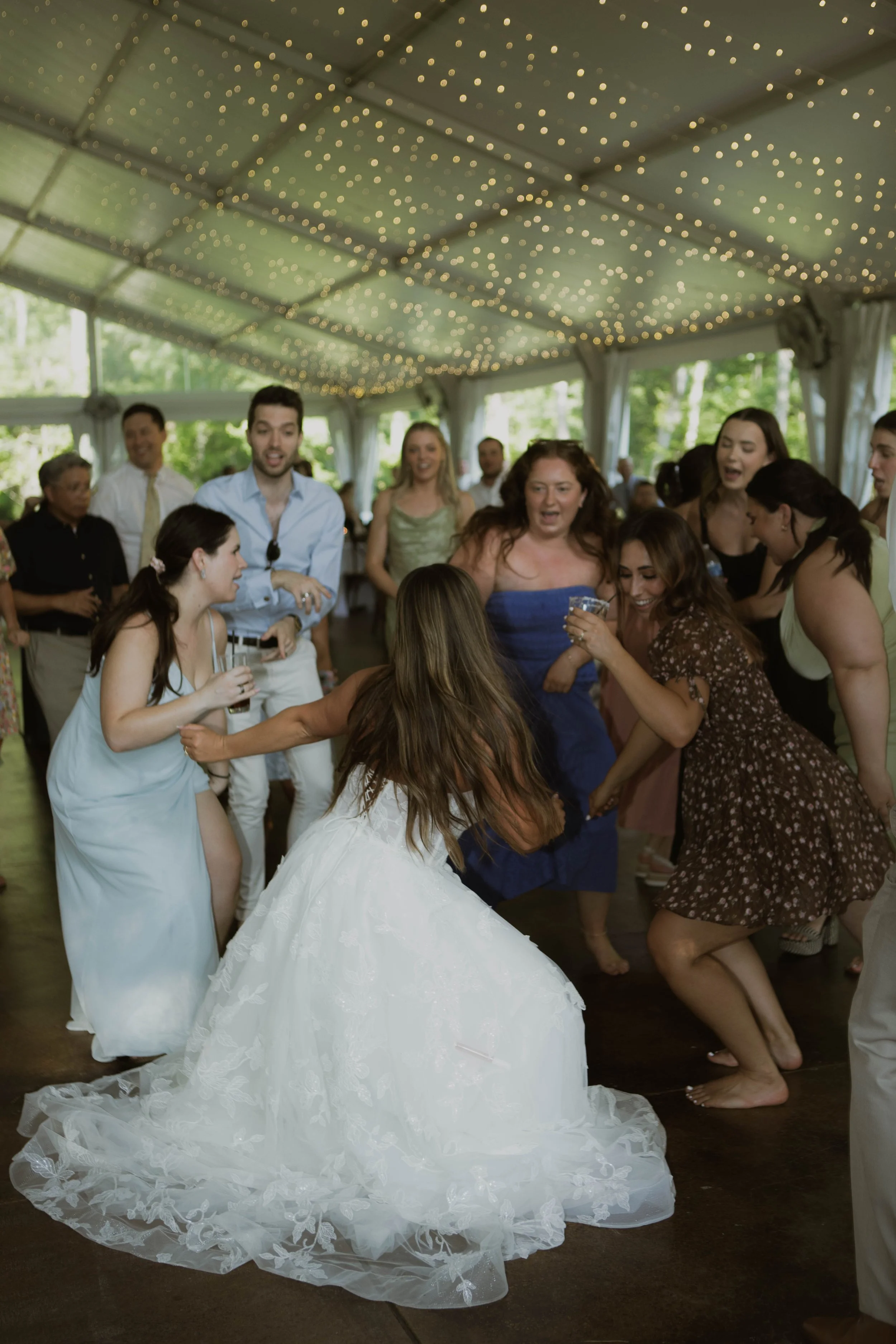 People dancing and celebrating at a wedding reception in a decorated tent with string lights.