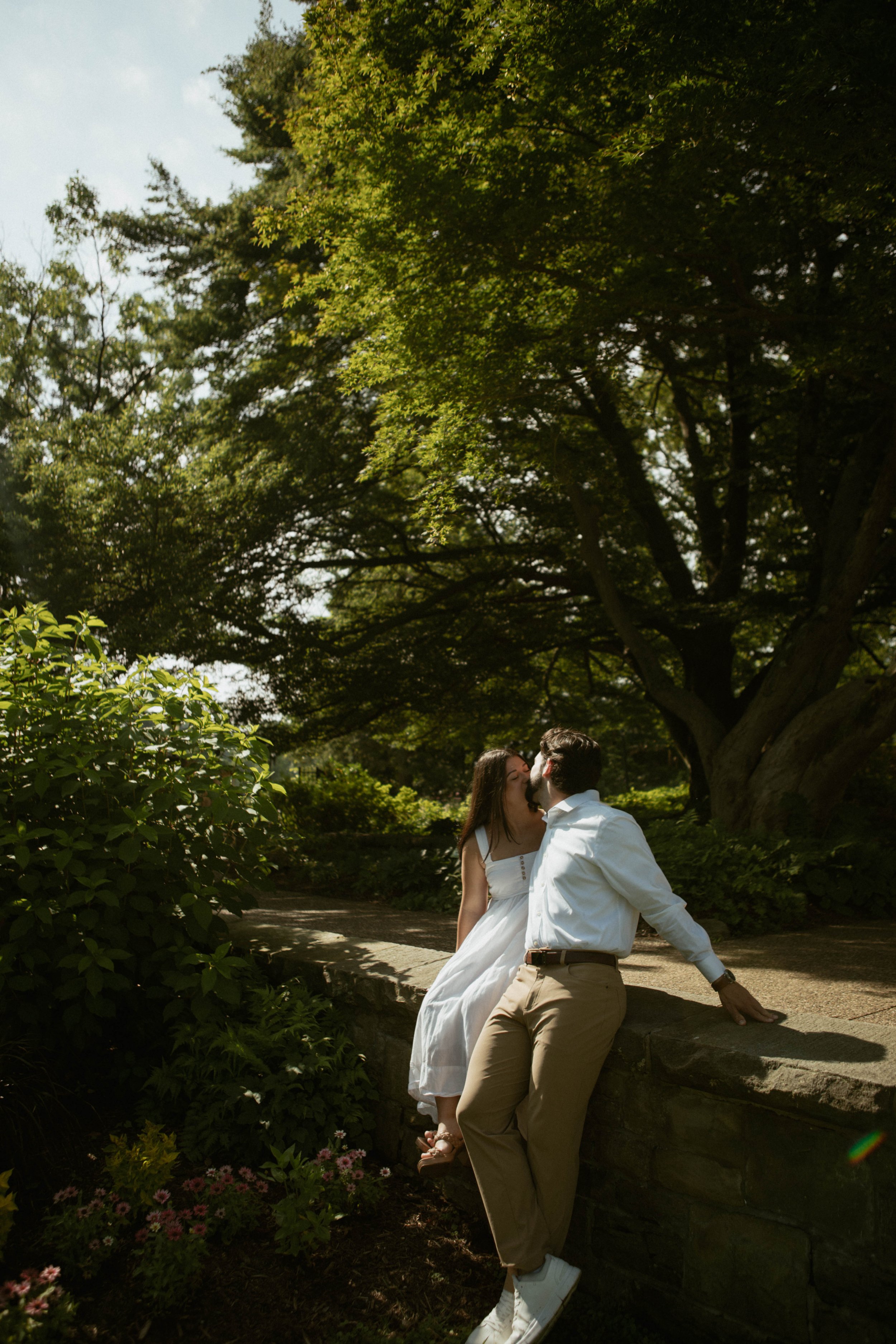 A couple sitting on a stone ledge in a park, sharing a kiss under large trees on a sunny day.