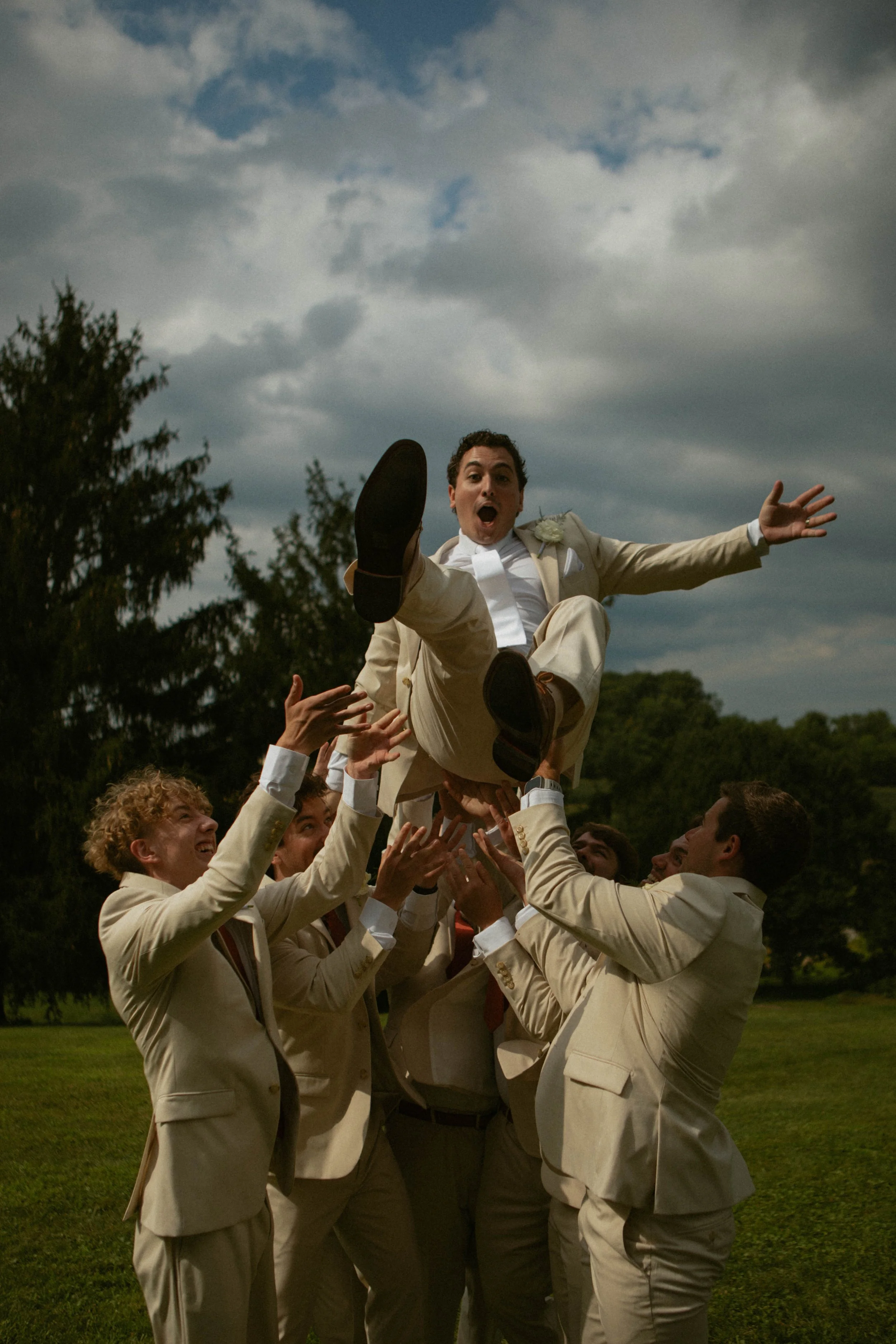 A groom in a beige suit being lifted into the air by groomsmen outdoors, with a cloudy sky and trees in the background.