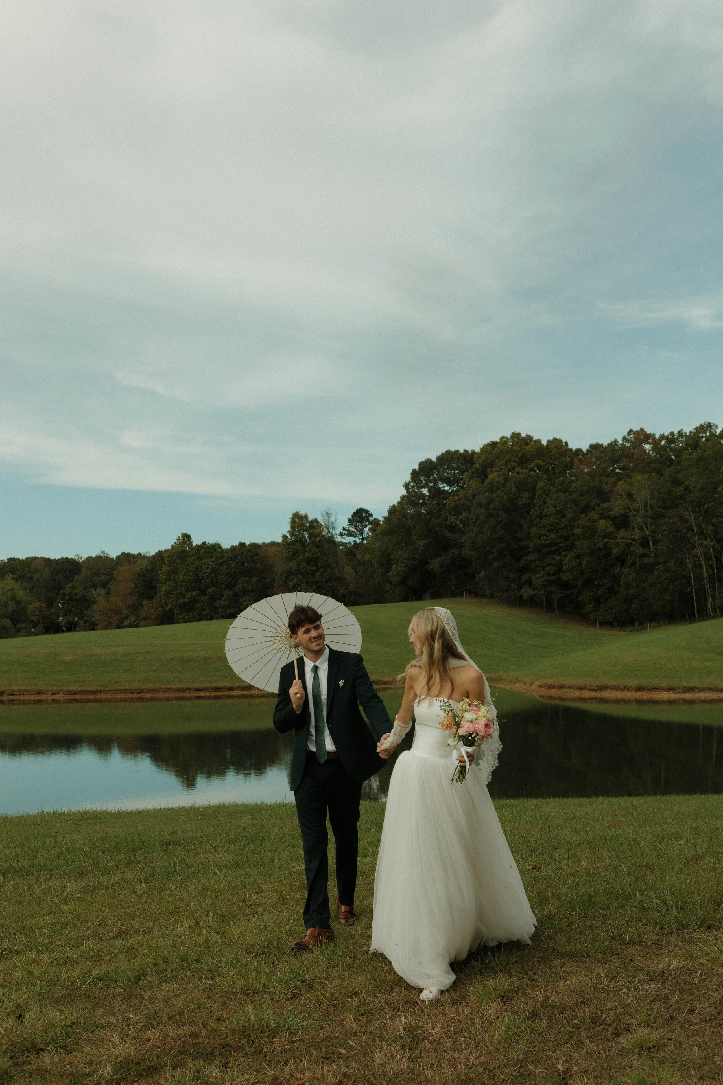 A couple in wedding attire walking by a lake, holding hands, with the groom holding an umbrella, outdoors in a scenic grassy area with trees and a cloudy sky.