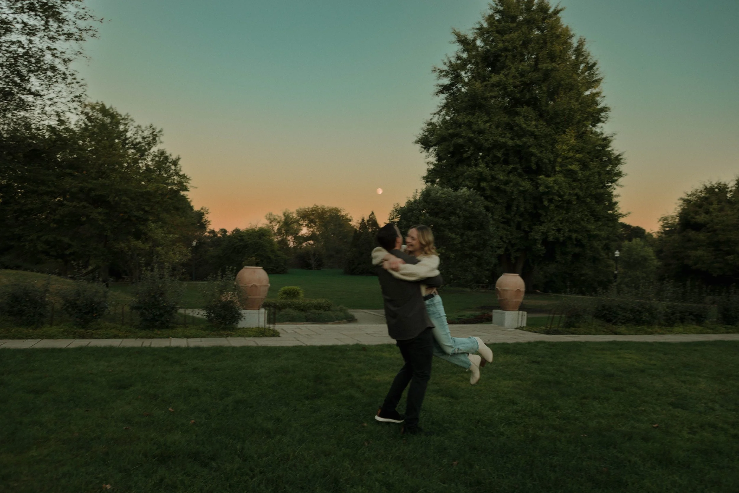 A couple is outdoors in a park at sunset, with the man lifting the woman in his arms. The woman is smiling and wearing light-colored jeans and a jacket, while the man is dressed in dark clothing. There are large decorative pots and trees in the backg