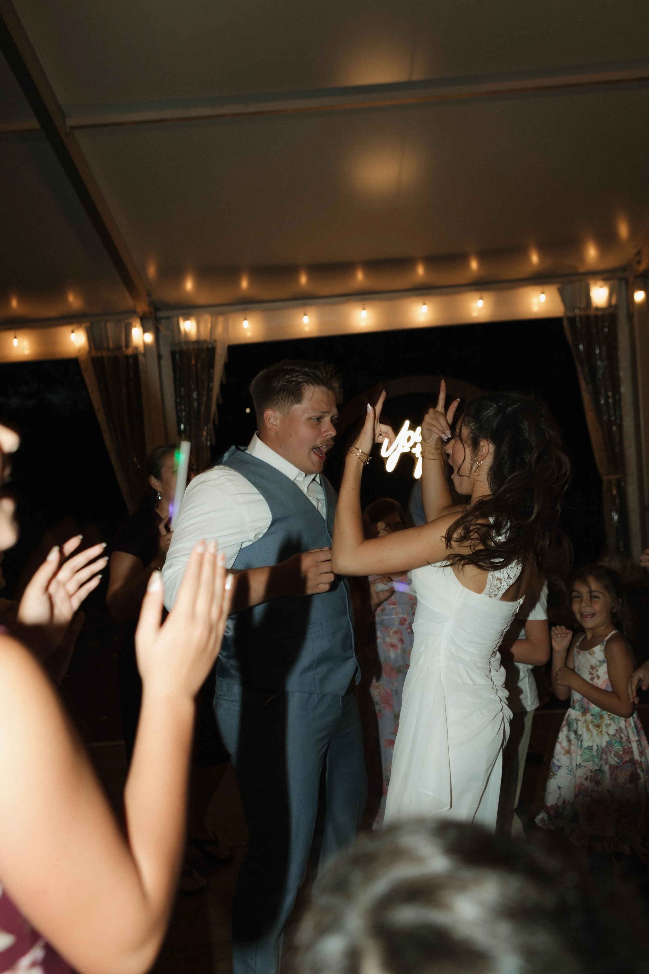 People dancing and celebrating at a wedding reception, with a bride and groom in the center, surrounded by family and friends under string lights.