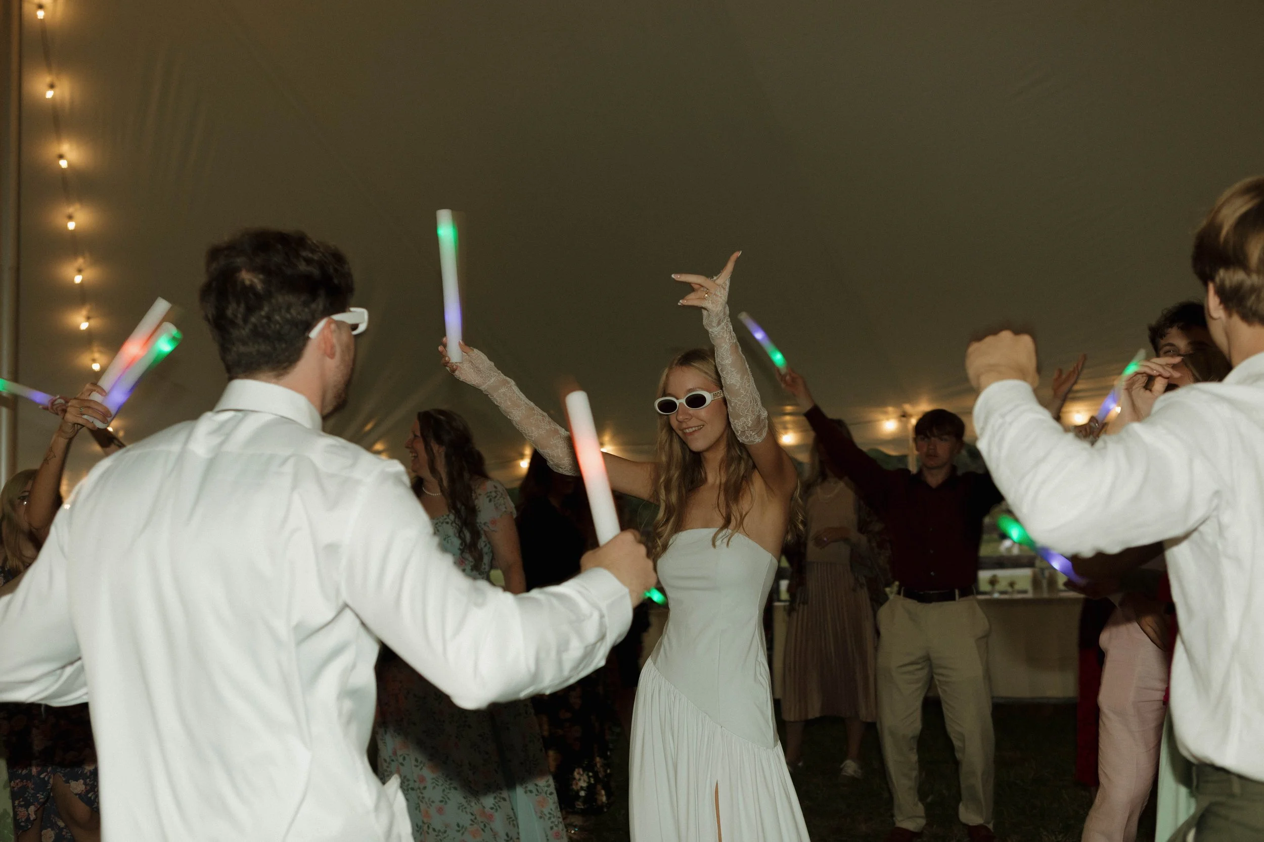 People dancing at a celebration under a tent, with a woman in a white dress and sunglasses dancing among others holding glow sticks.