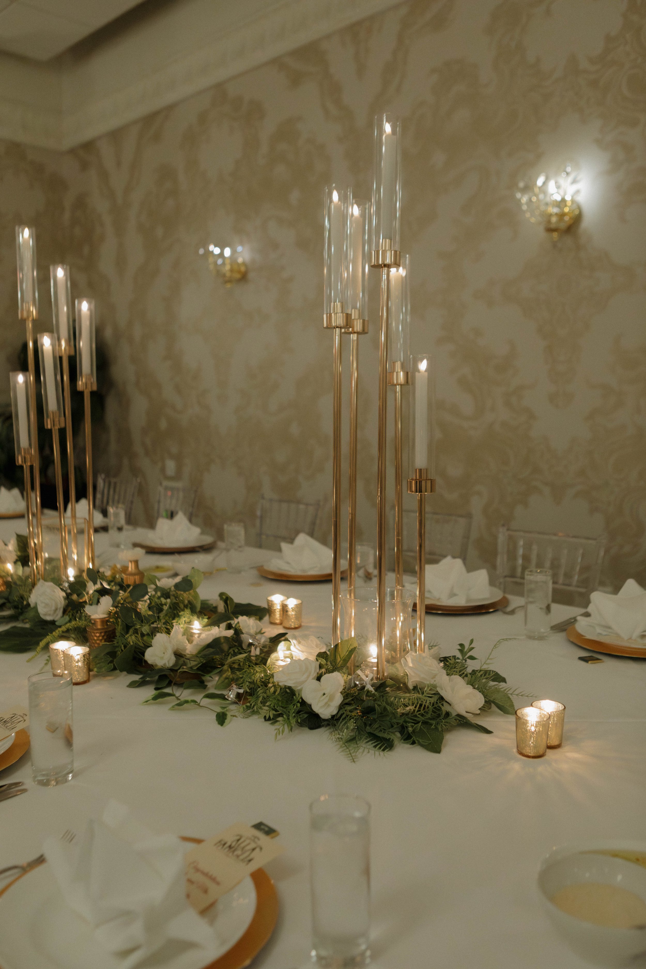 Elegant banquet table with white tablecloth, floral centerpiece, tall gold candle holders with white candles, small gold votive candles, with place settings including white napkins and water glasses, in a decorated room with patterned wallpaper.