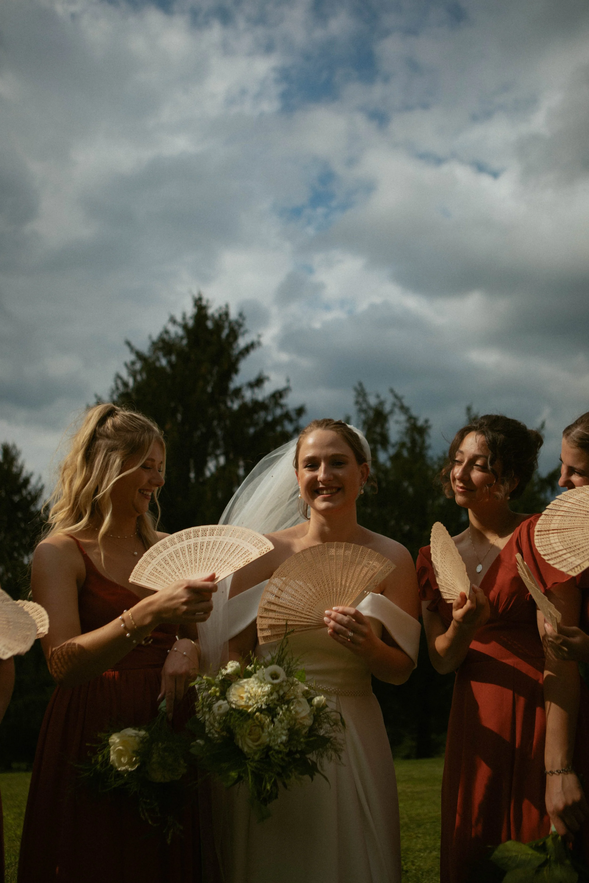 A bride in a white wedding dress holding a bouquet of white flowers, surrounded by bridesmaids in matching reddish-brown dresses, all holding hand fans, outdoors under a cloudy sky.