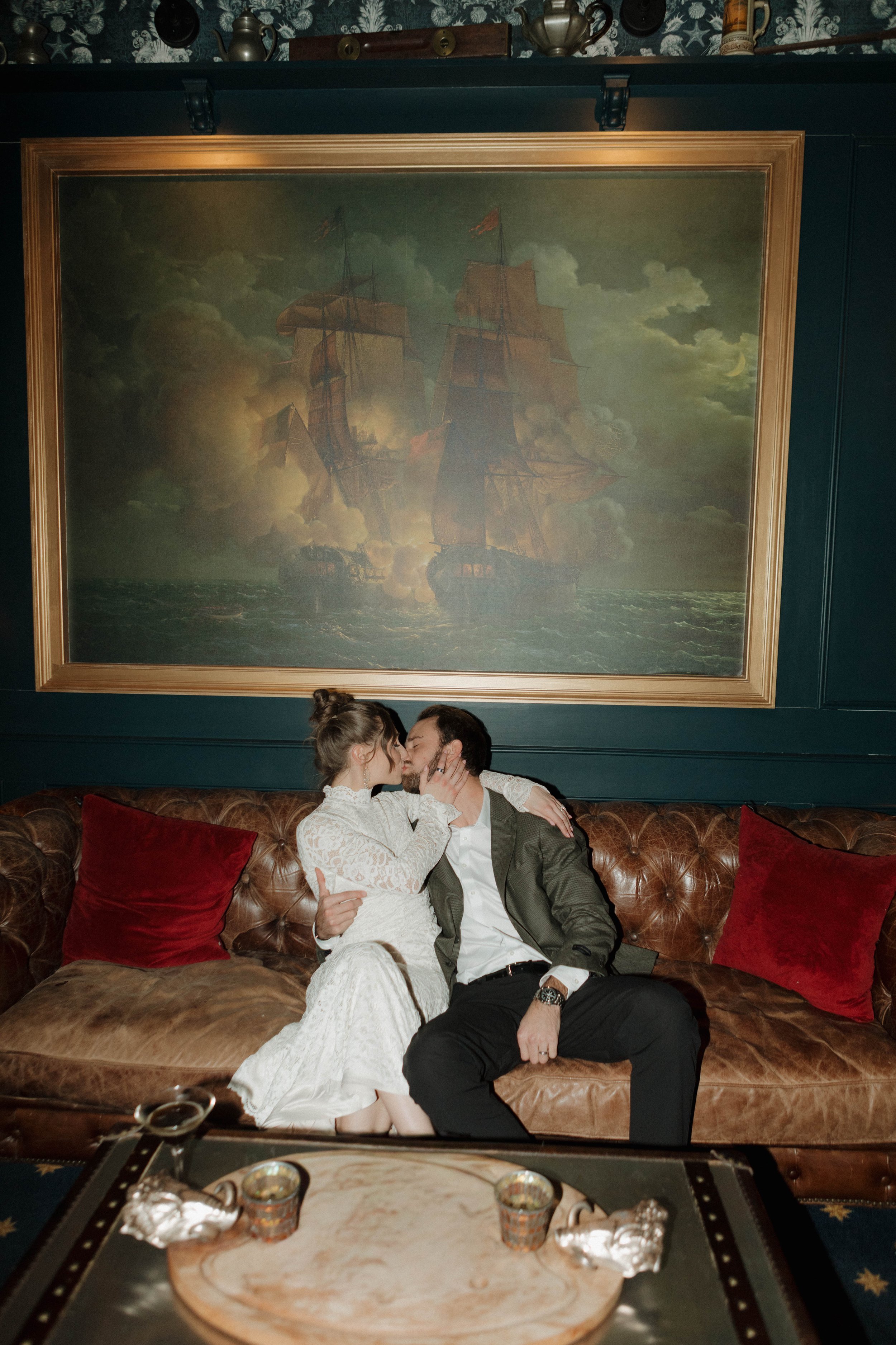 A couple sits on a brown leather sofa in a dimly lit room, sharing a kiss. The woman is wearing a white lace dress, and the man is in a dark suit with a white shirt. Behind them is a large framed nautical painting of sailing ships on the sea, with dark clouds in the sky. There are red pillows on the sofa, and a coffee table with small cups and decorative items in front of them.