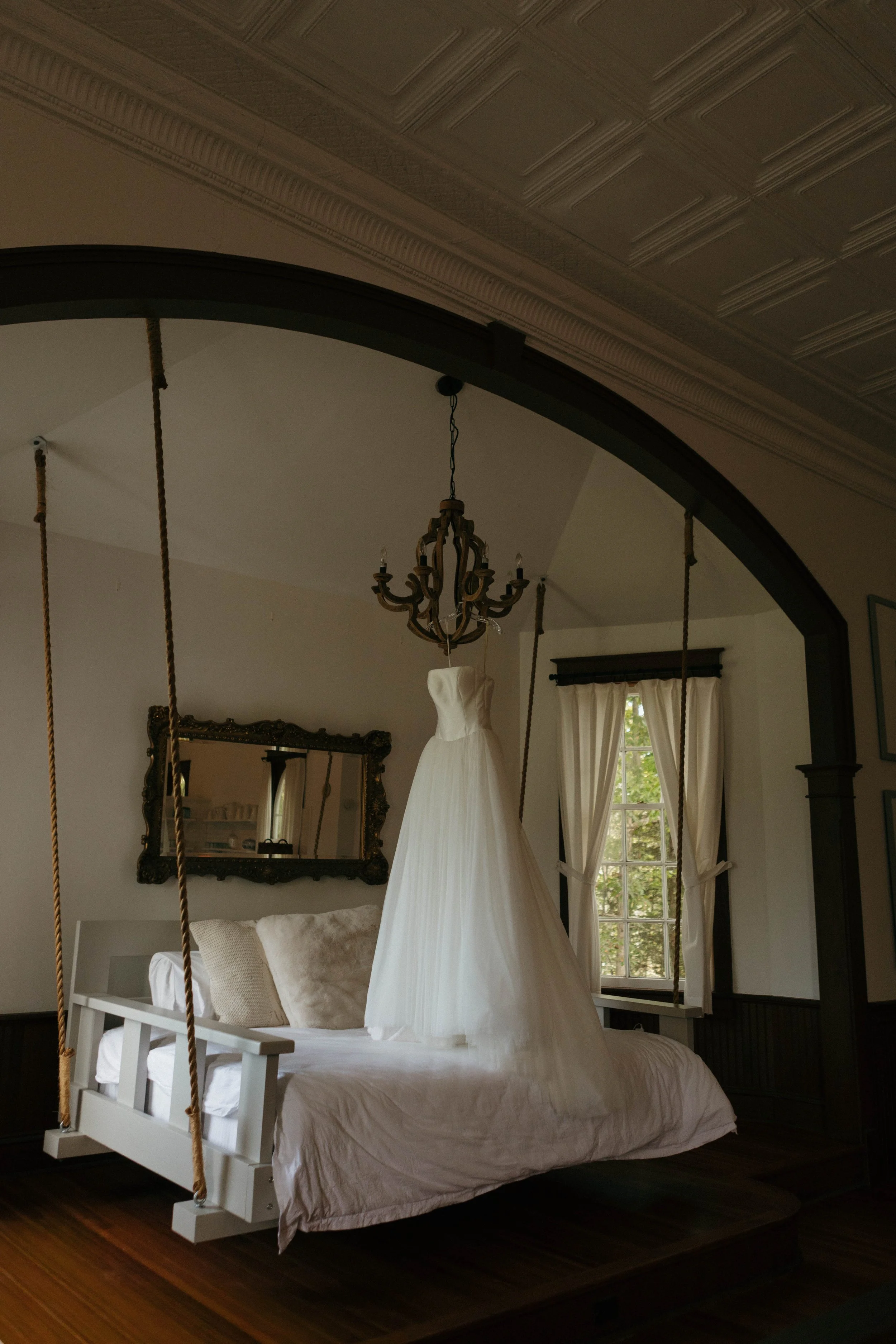 White wedding dress hanging from a chandelier above a bed with white linens in a room with a window and curtains.