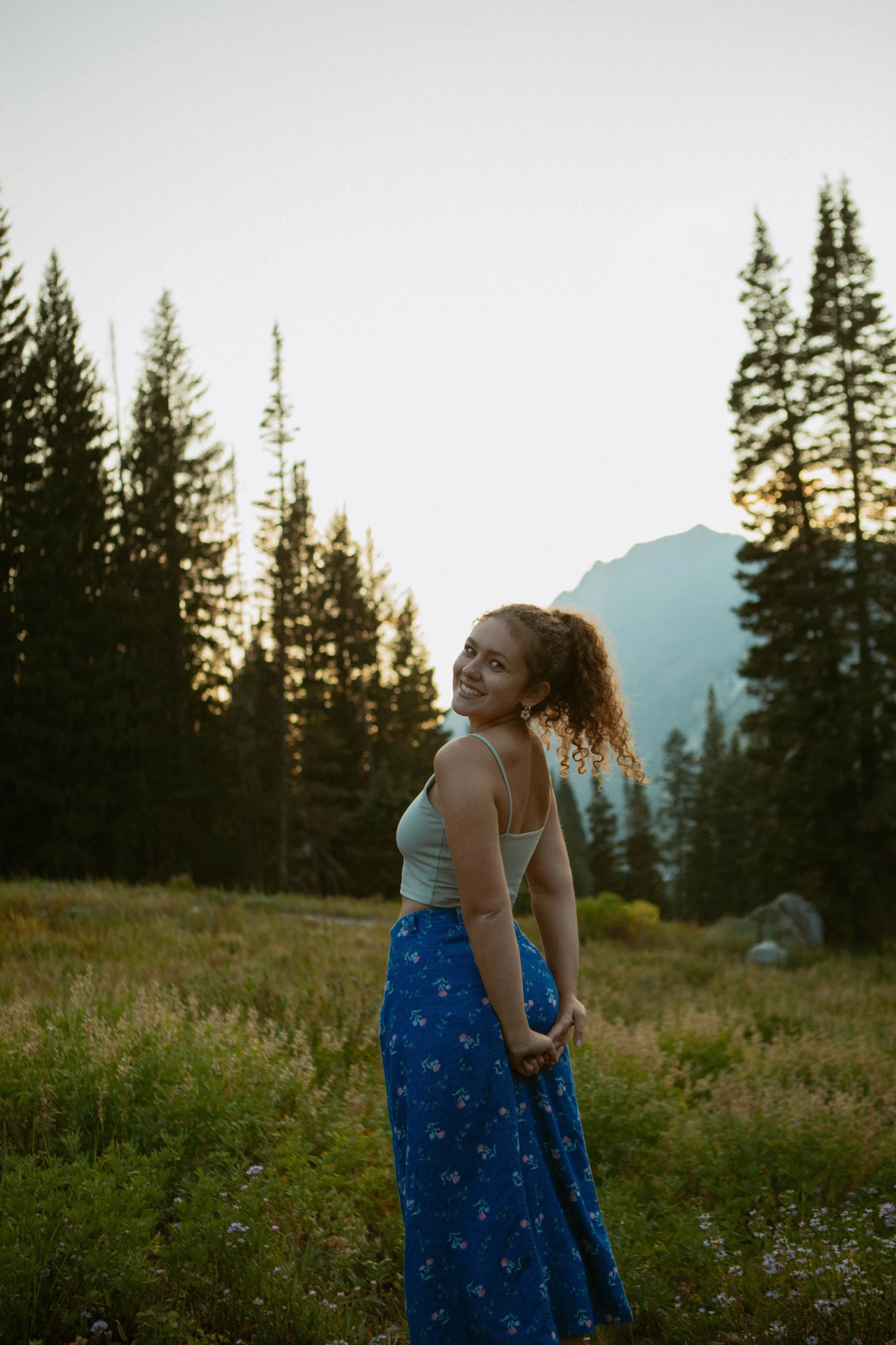 A young woman with curly hair standing in a field at sunset, surrounded by trees, smiling and looking at the camera.