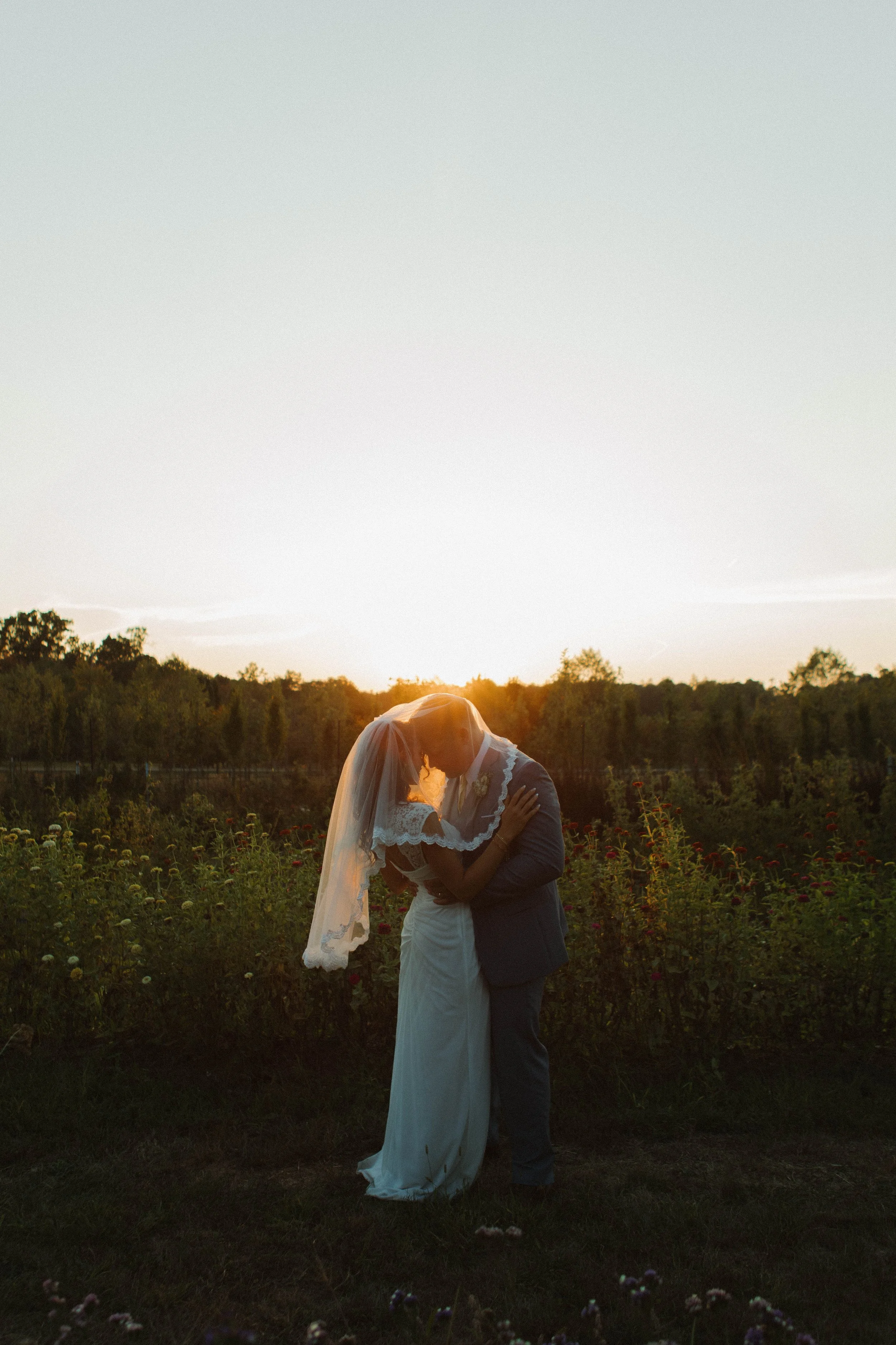 A couple dressed in wedding attire sharing a kiss outdoors at sunset.