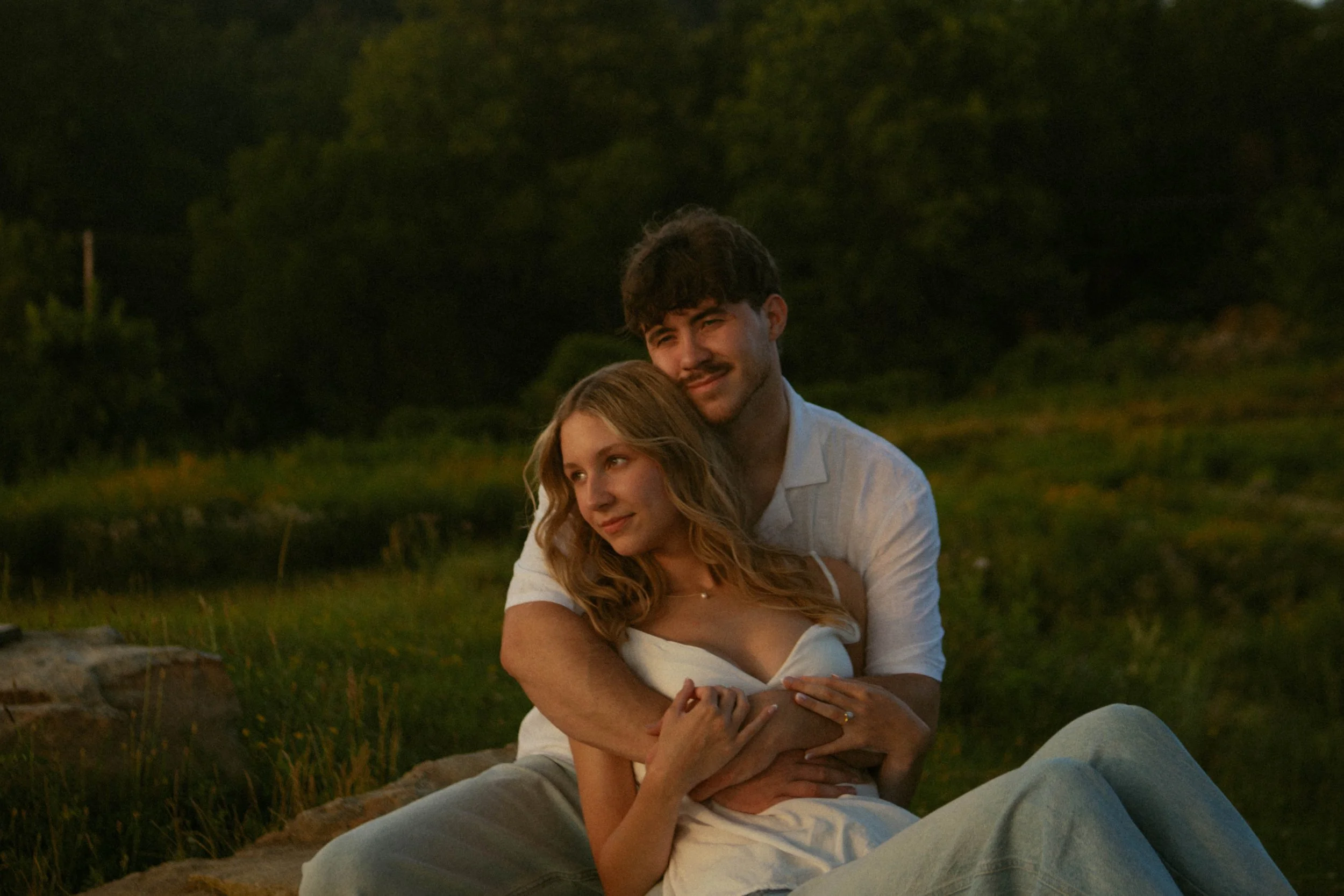 A young couple sitting outdoors on a rock during sunset, with the man hugging the woman from behind as they look contentedly into the distance.