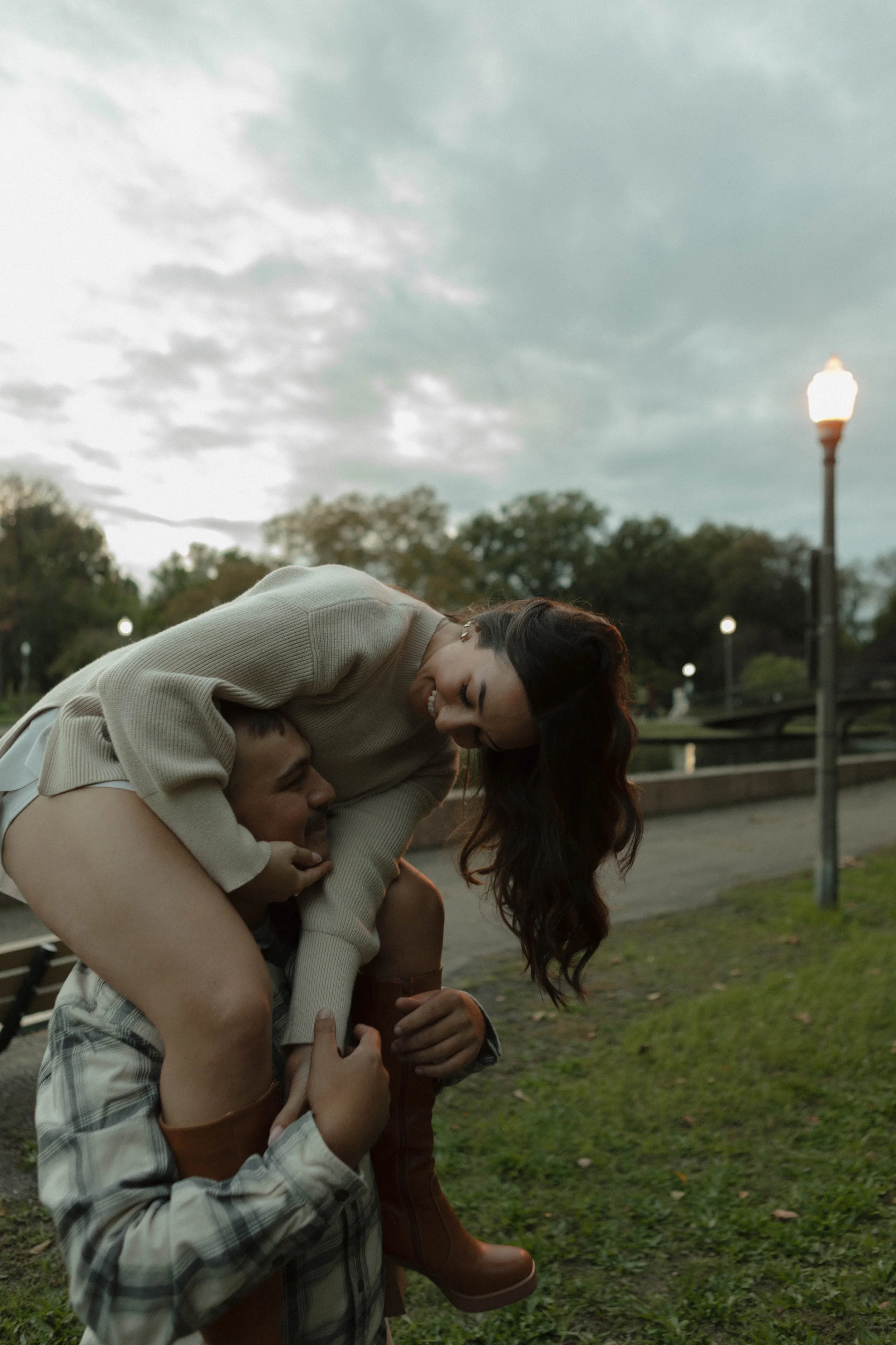 A man carrying a woman on his shoulders in a park during early evening with streetlights on.