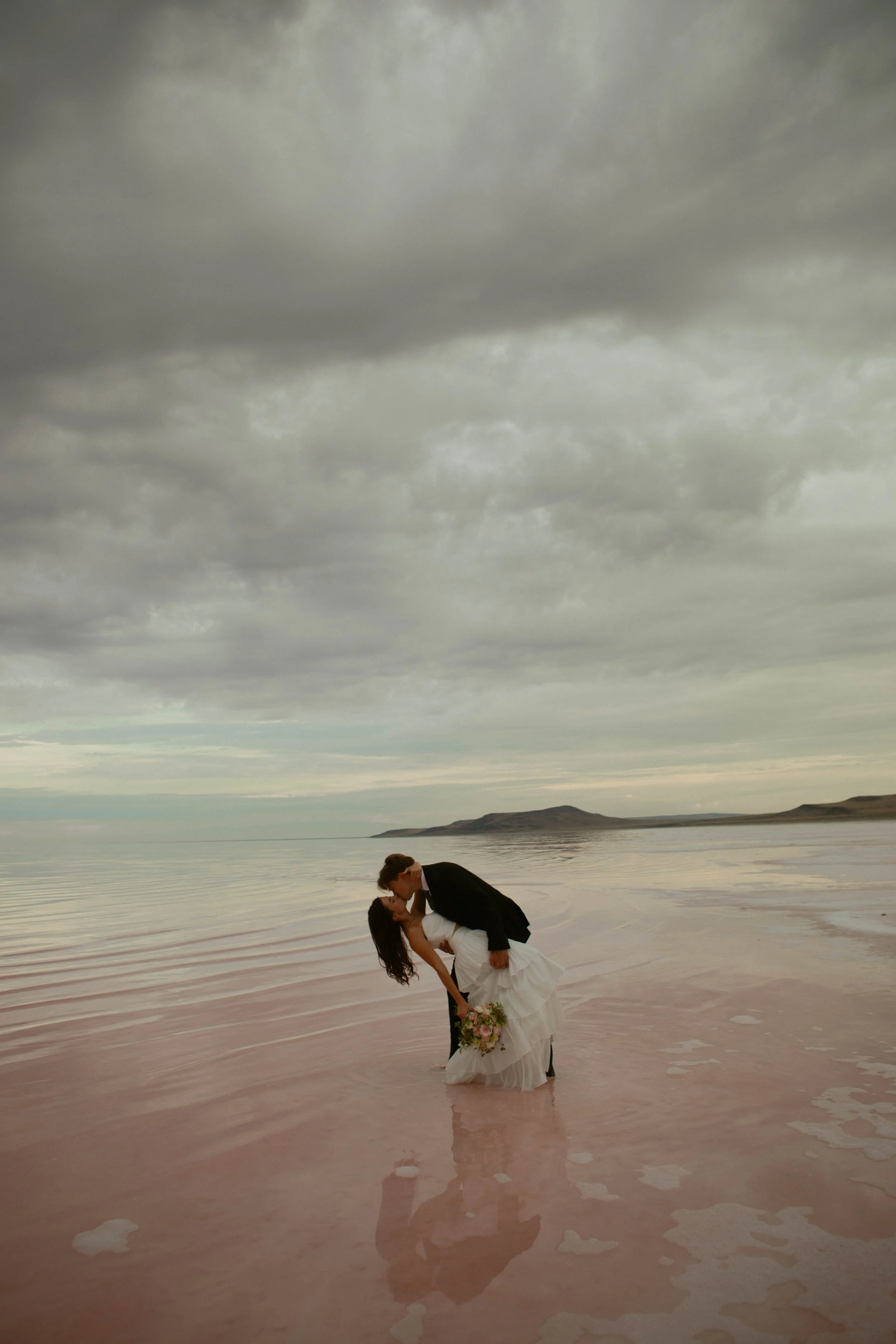 A couple in wedding attire sharing a kiss in shallow pink water on a beach under cloudy sky.