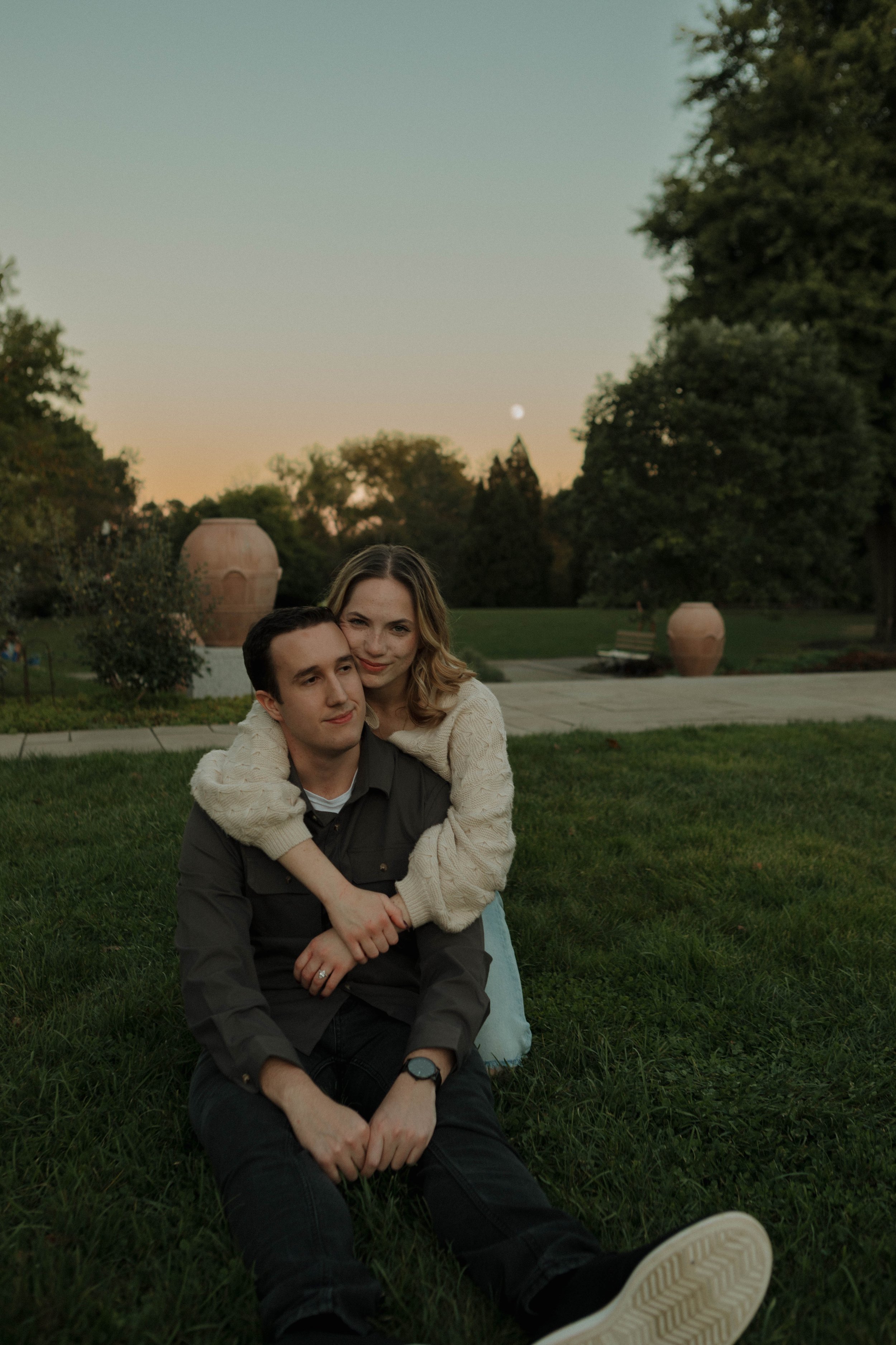 A young couple sitting on the grass in a park at dusk, with the woman hugging the man from behind. The sky is a gradient of soft colors with a visible moon, large trees, and large decorative pots in the background.
