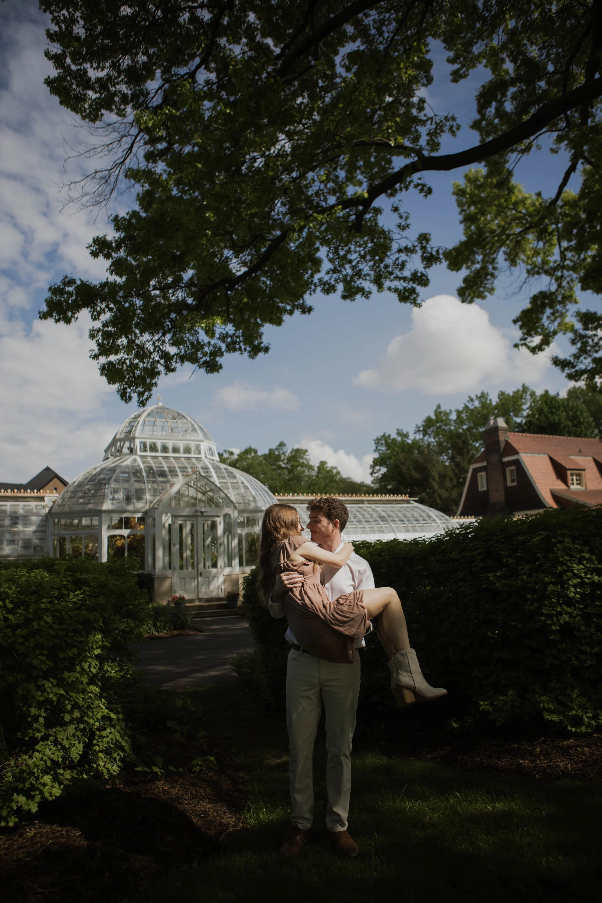 A man carrying a woman in his arms outdoors near a glass greenhouse, with trees and a blue sky with clouds in the background.