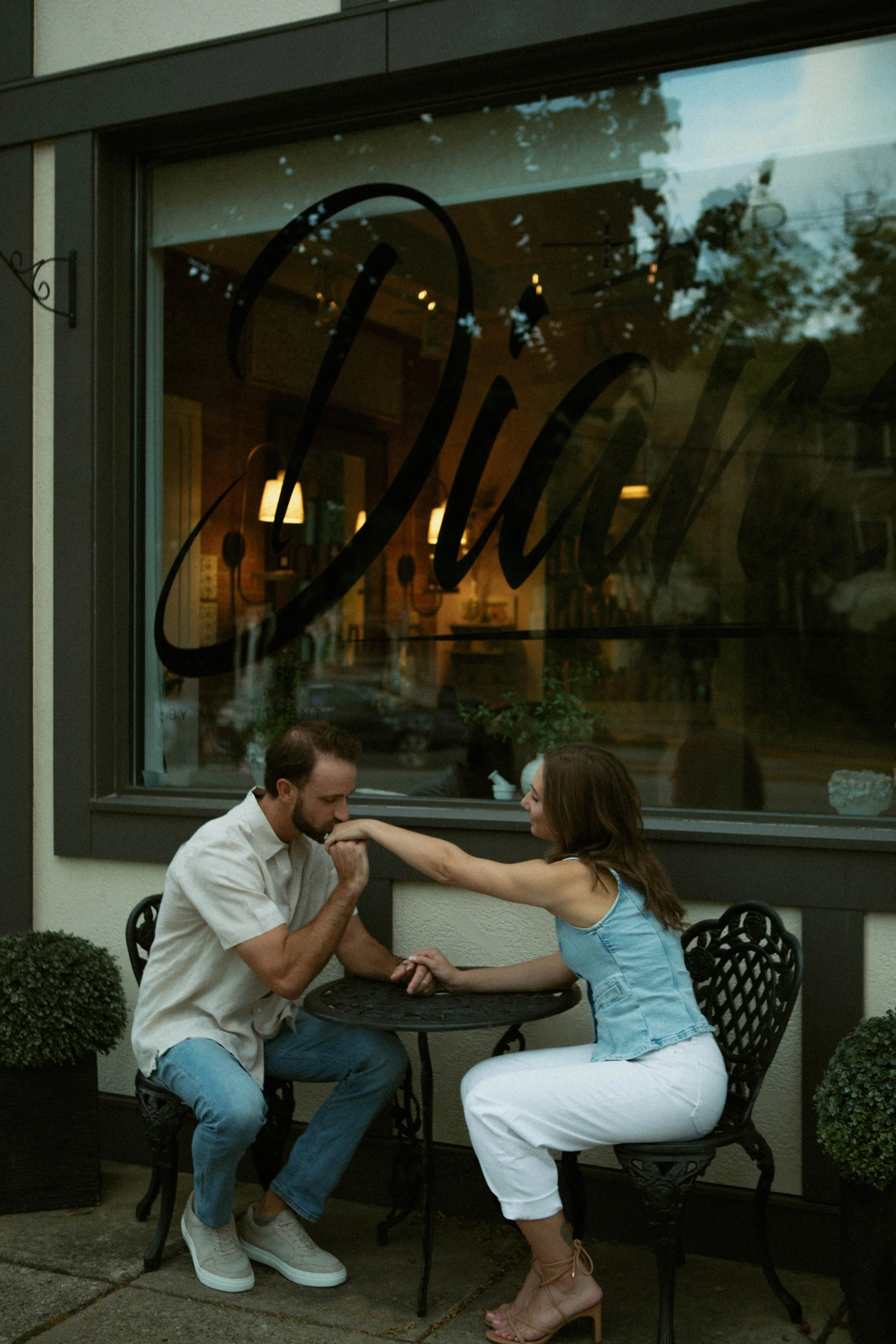 A couple sitting outside a café, with the man kissing the woman's arm. The scene appears intimate and affectionate.