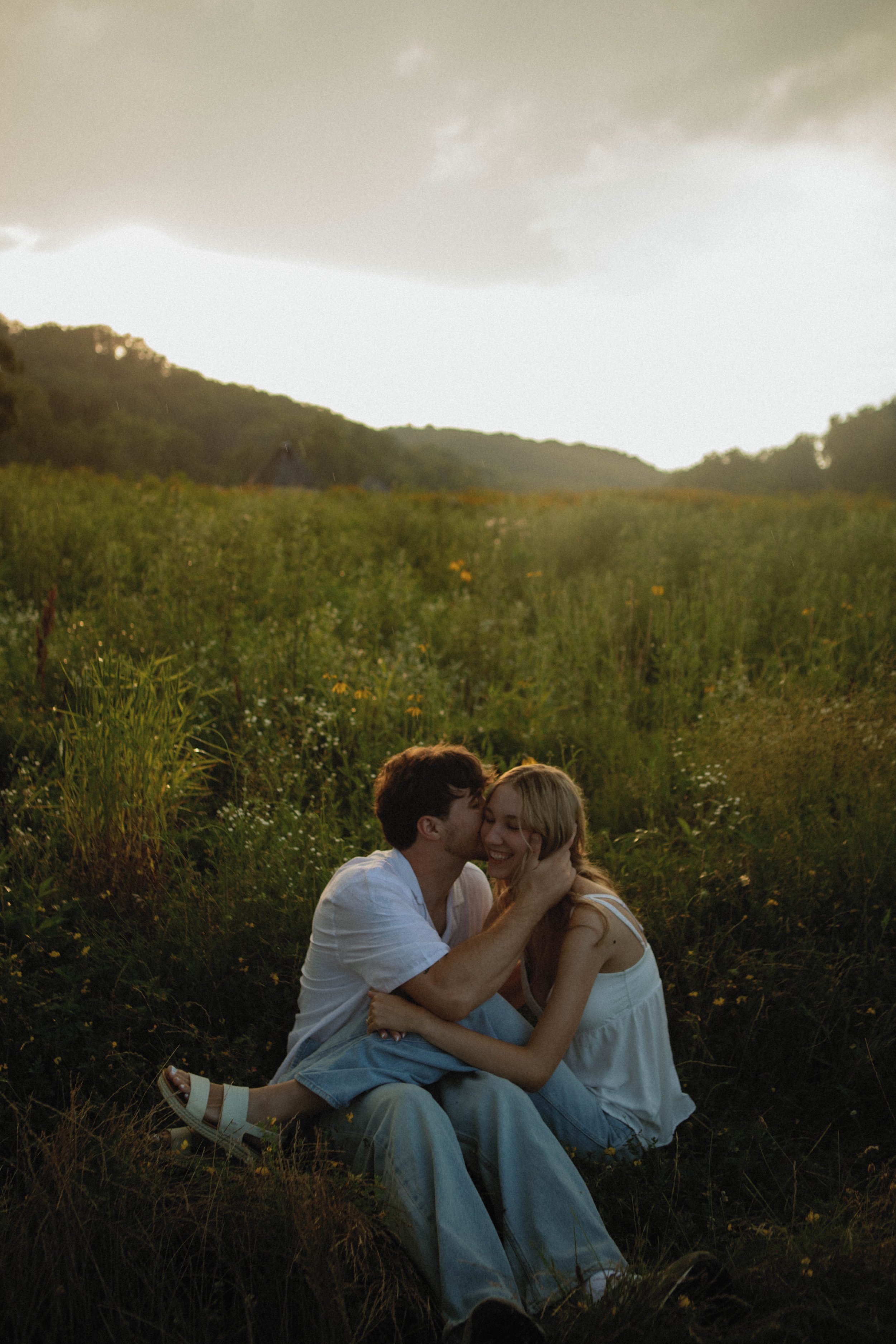 A couple sitting on the grass in a field hugging and smiling at each other during sunset.