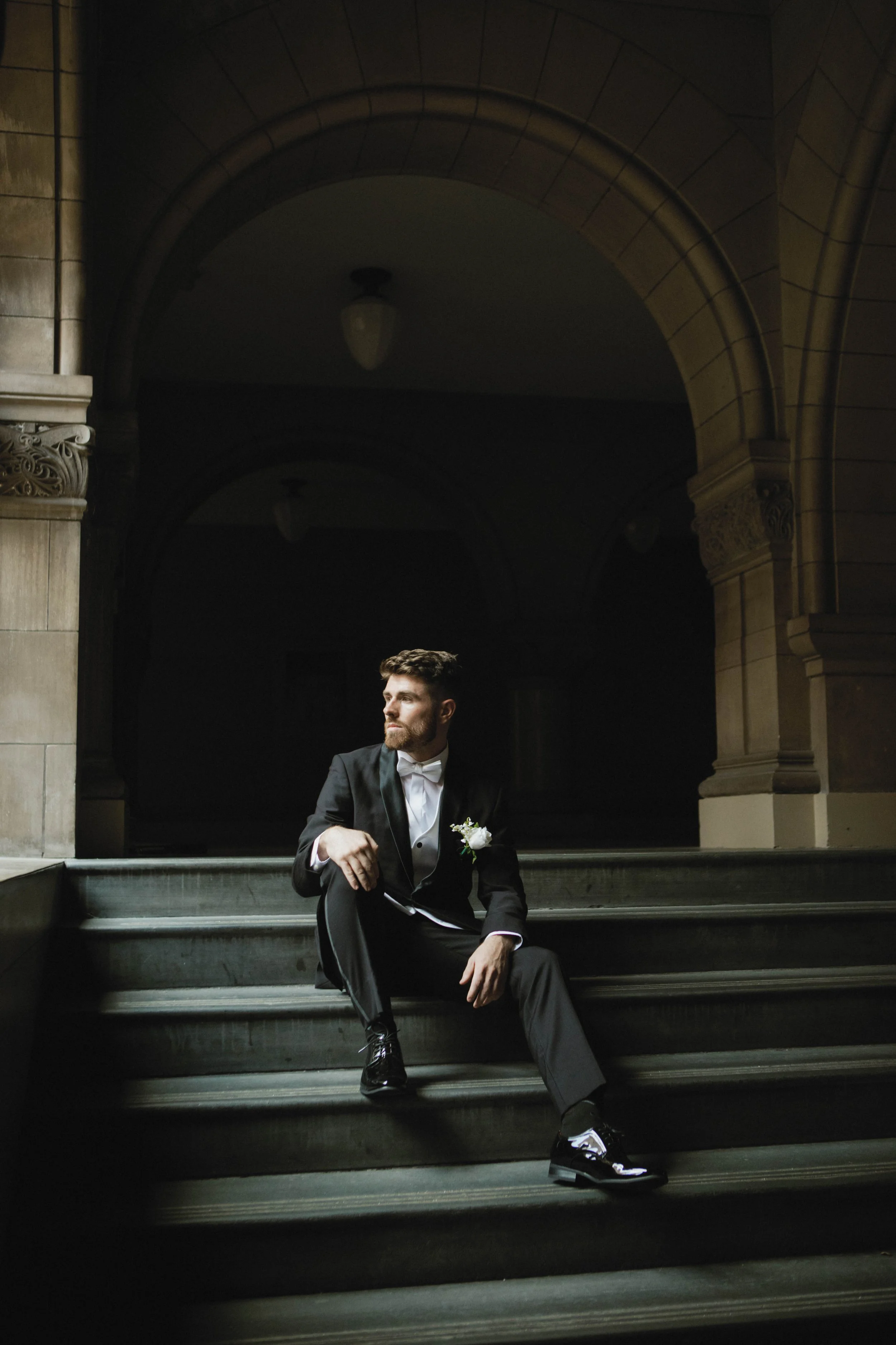 A man in a black tuxedo with a white shirt and bowtie, sitting on stone stairs in an architectural setting with arches and columns, holding a cane and wearing a white boutonniere.