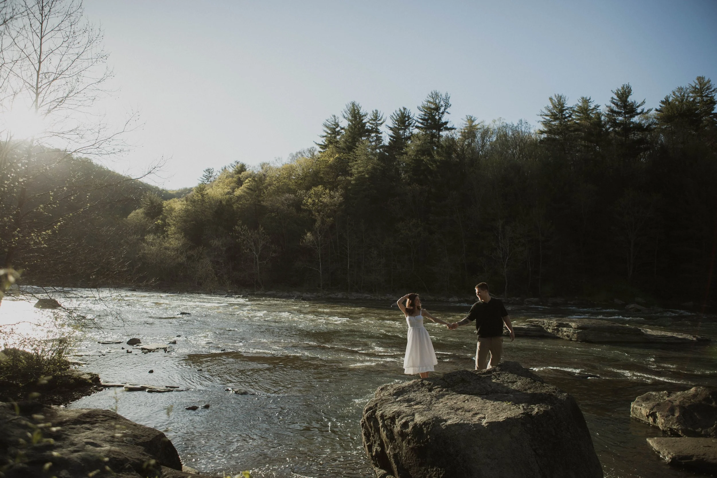A couple holding hands in a flowing river surrounded by trees and rocks, with sunlight shining on the scene.