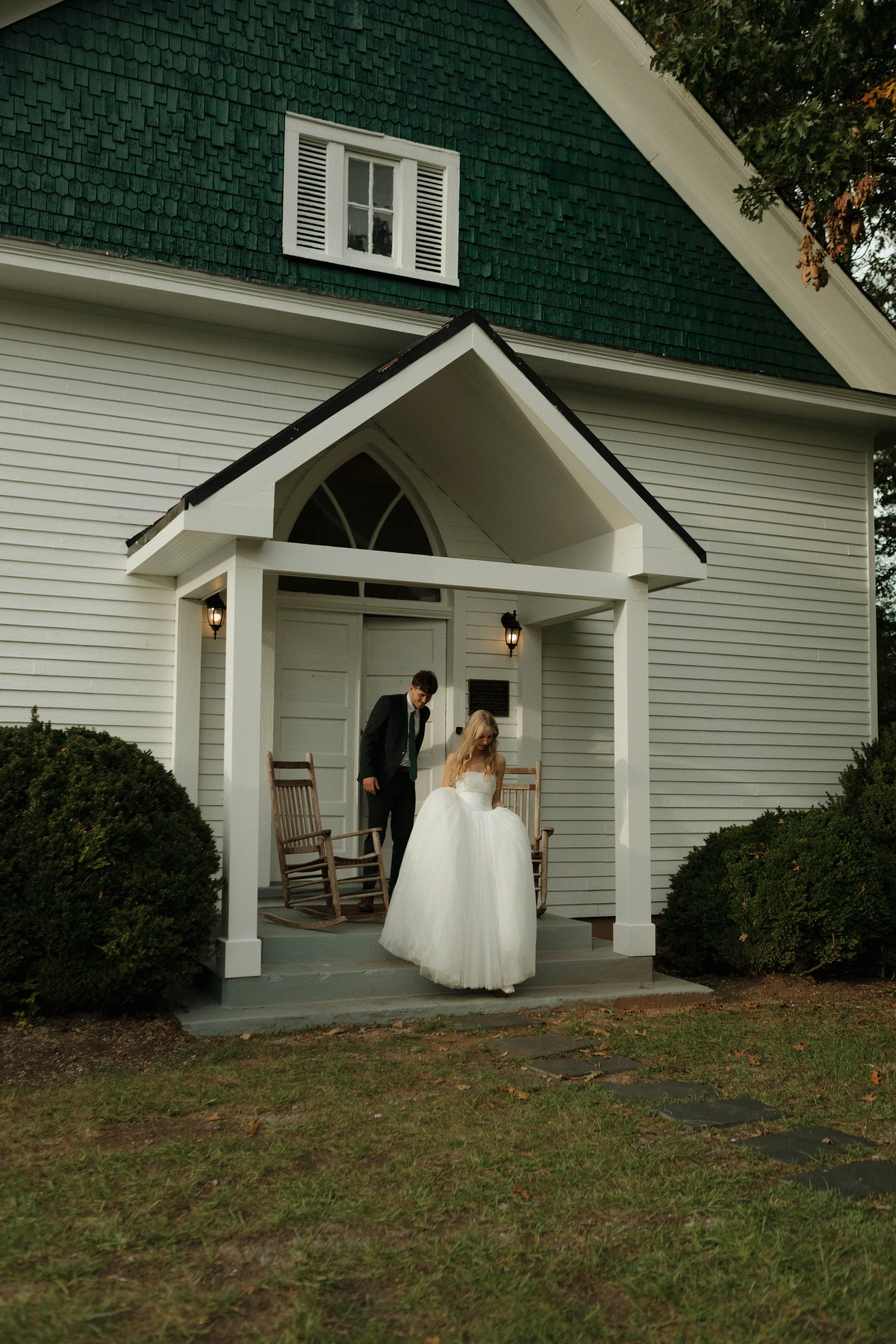A bride in a white wedding dress and a groom in a black suit standing on the front porch of a white house with a green roof. The house has a small porch with two rocking chairs. The bride is looking down, the groom is looking at her, and there are bushes on either side of the porch.