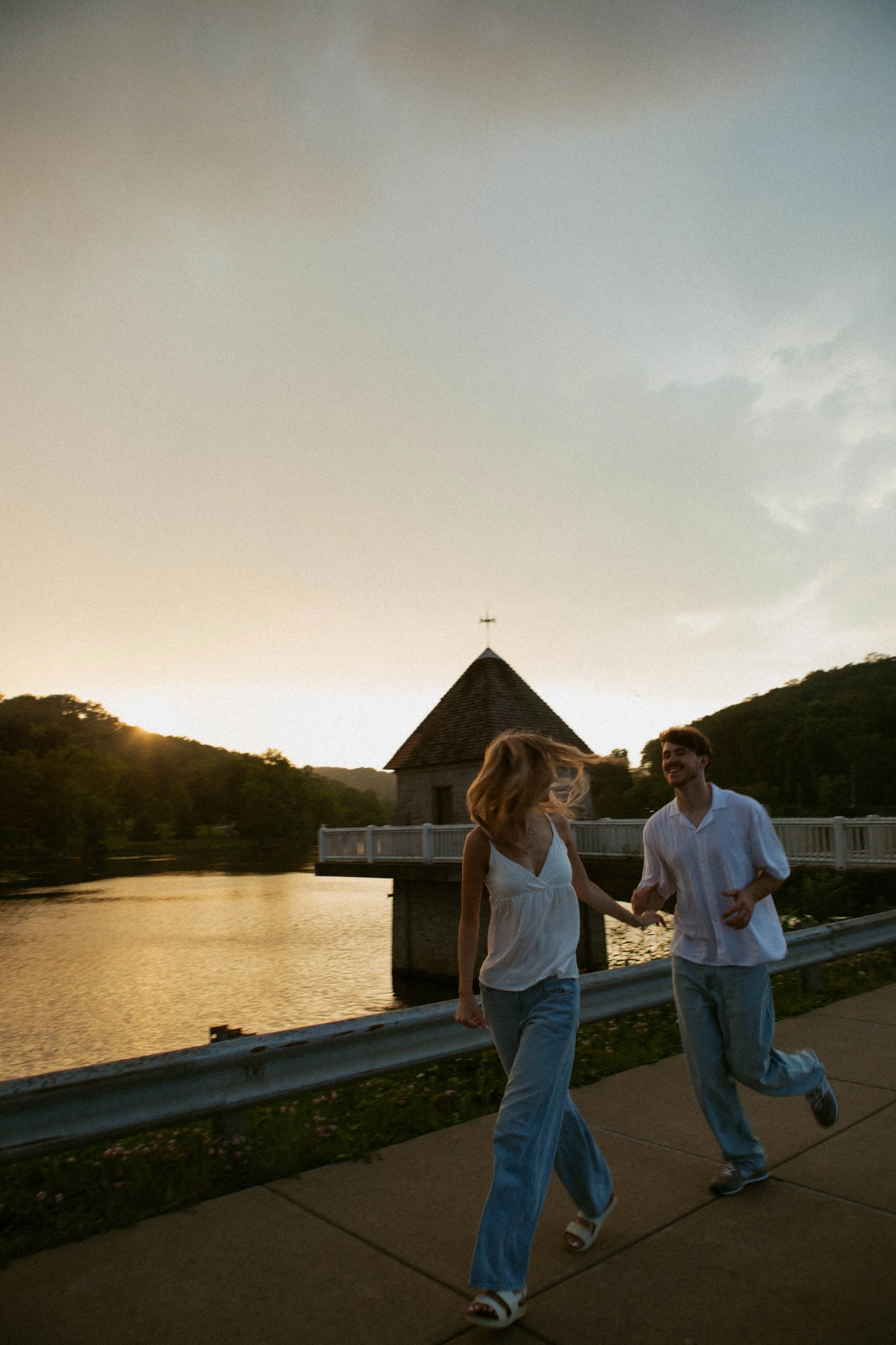 A couple holding hands and running along a sidewalk at sunset, with a body of water and a small church visible in the background.