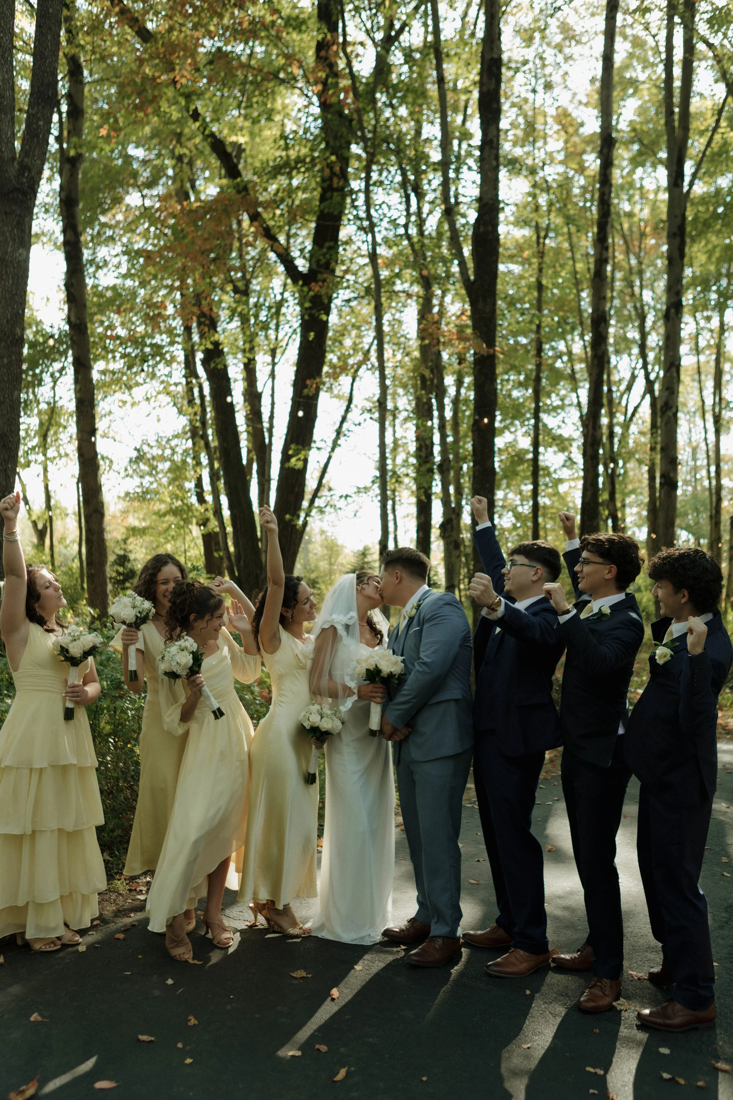 A group of bridesmaids and groomsmen, dressed in yellow and navy suits, standing outdoors on a paved path in a forest, celebrating a wedding. The bride and groom are kissing, surrounded by friends cheering.