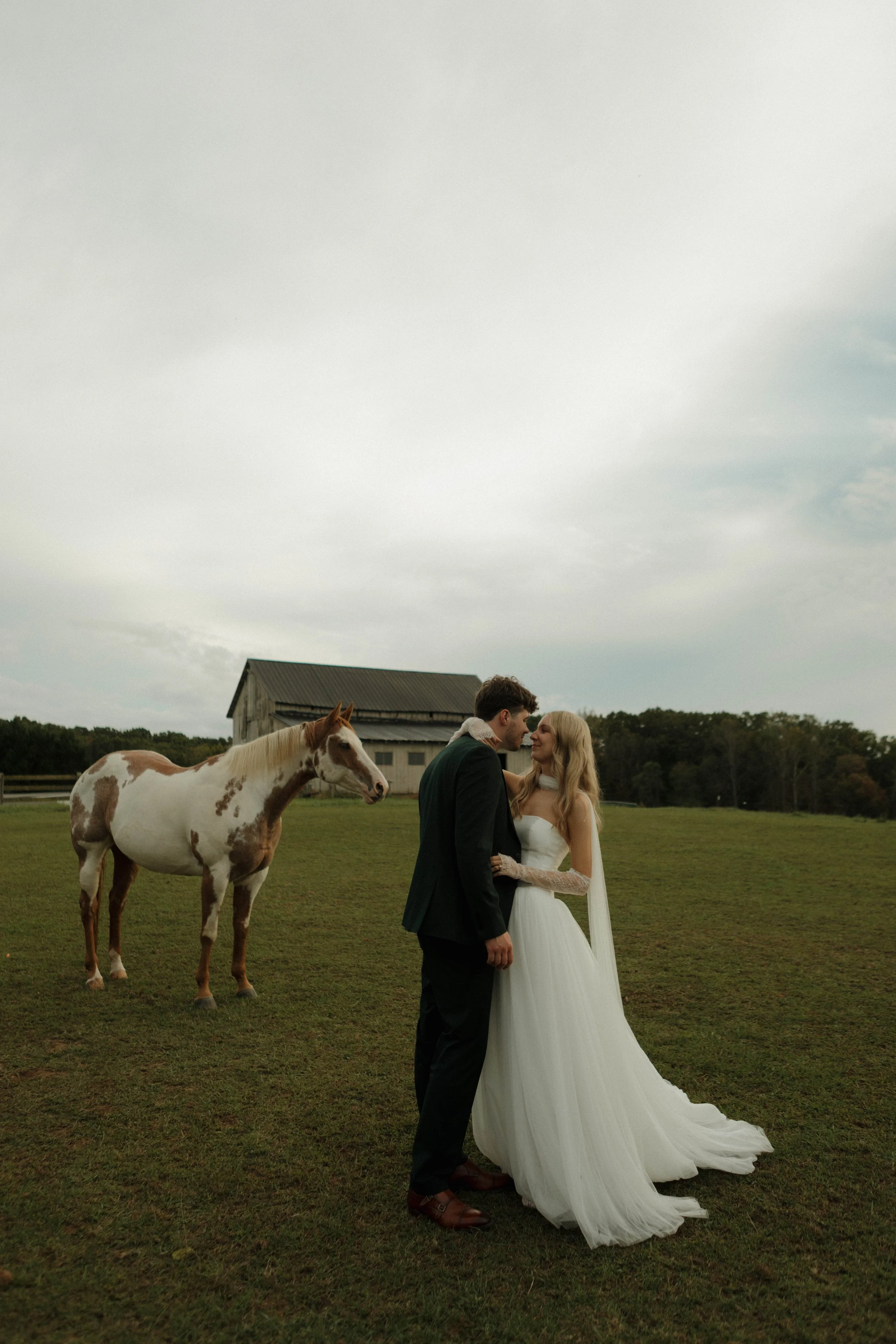 A bride and groom standing close together in a field, with a horse nearby and a barn in the background on a cloudy day.