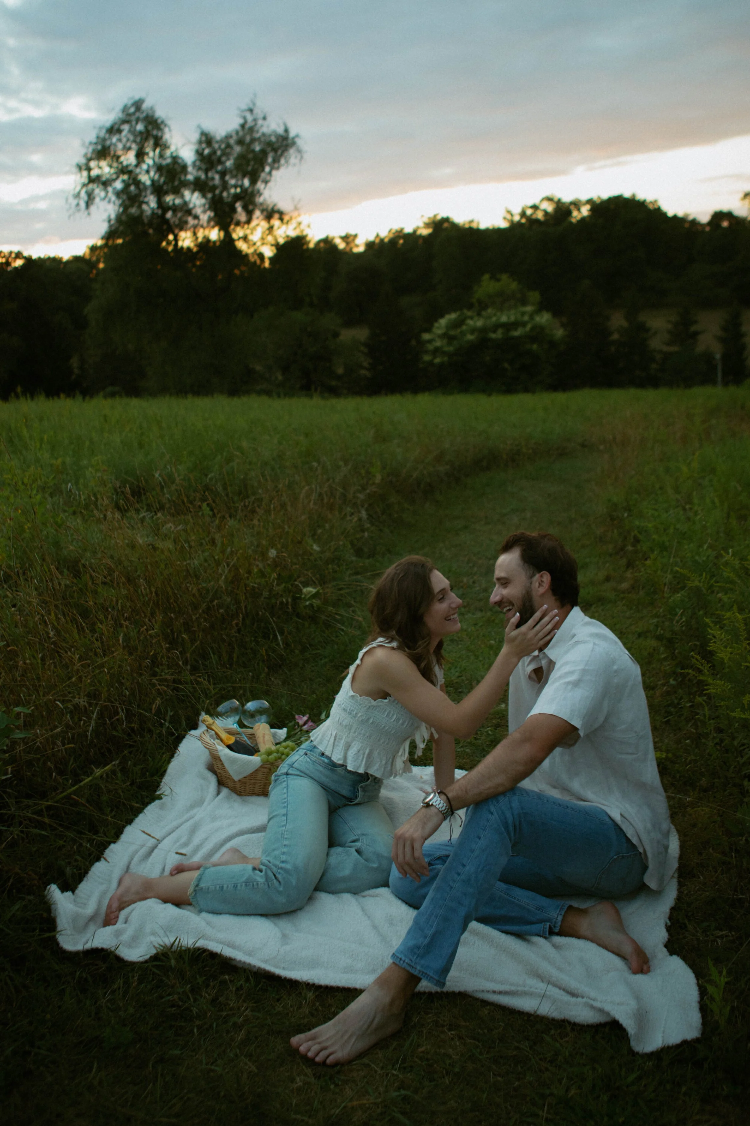 A young woman touches a man's face during a romantic picnic on a white blanket in a grassy field at sunset. There is a picnic basket with food and drinks nearby, and trees and hills are visible in the background.