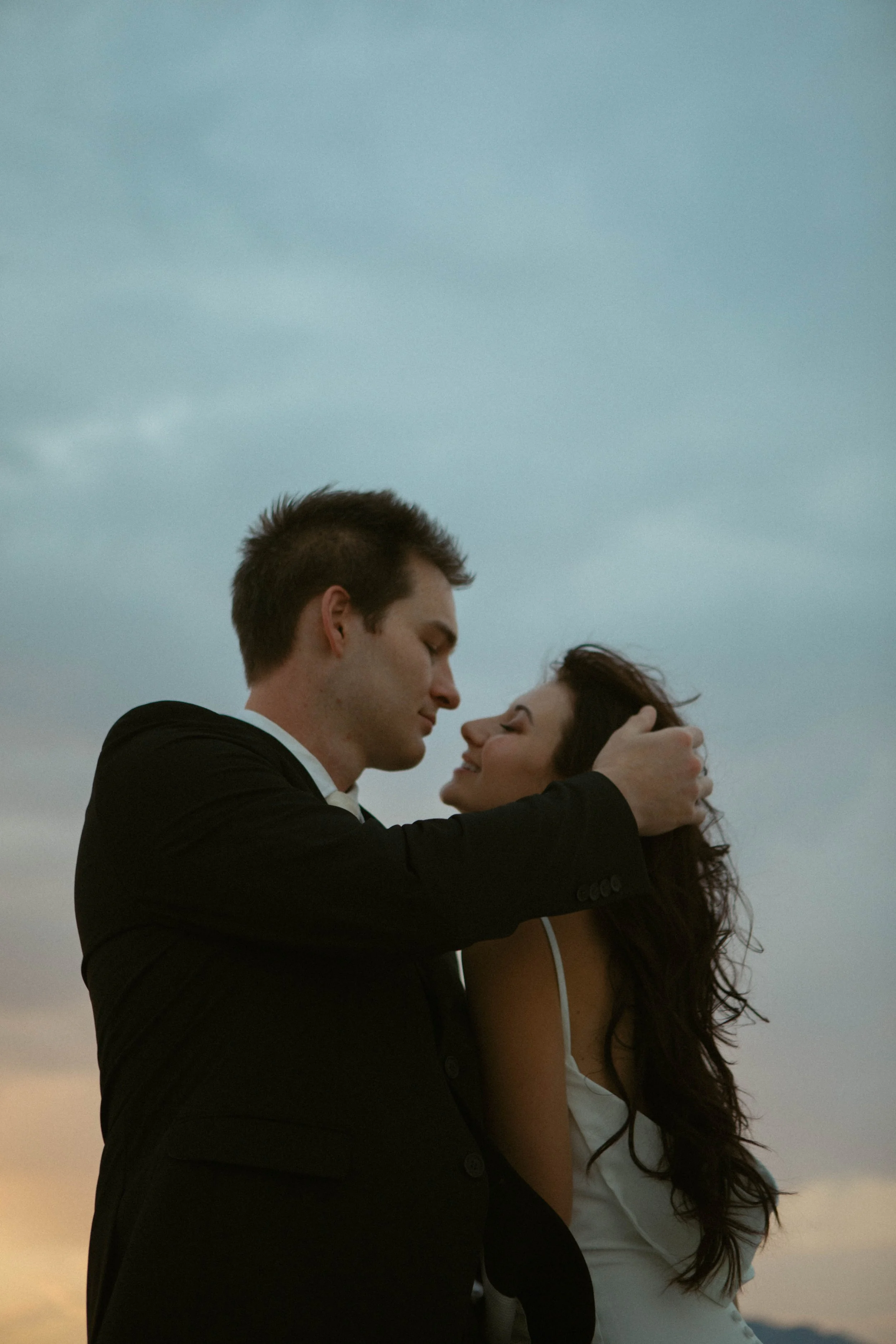 A romantic couple sharing an intimate moment outdoors at sunset, with the man holding the woman's face, both smiling gently.