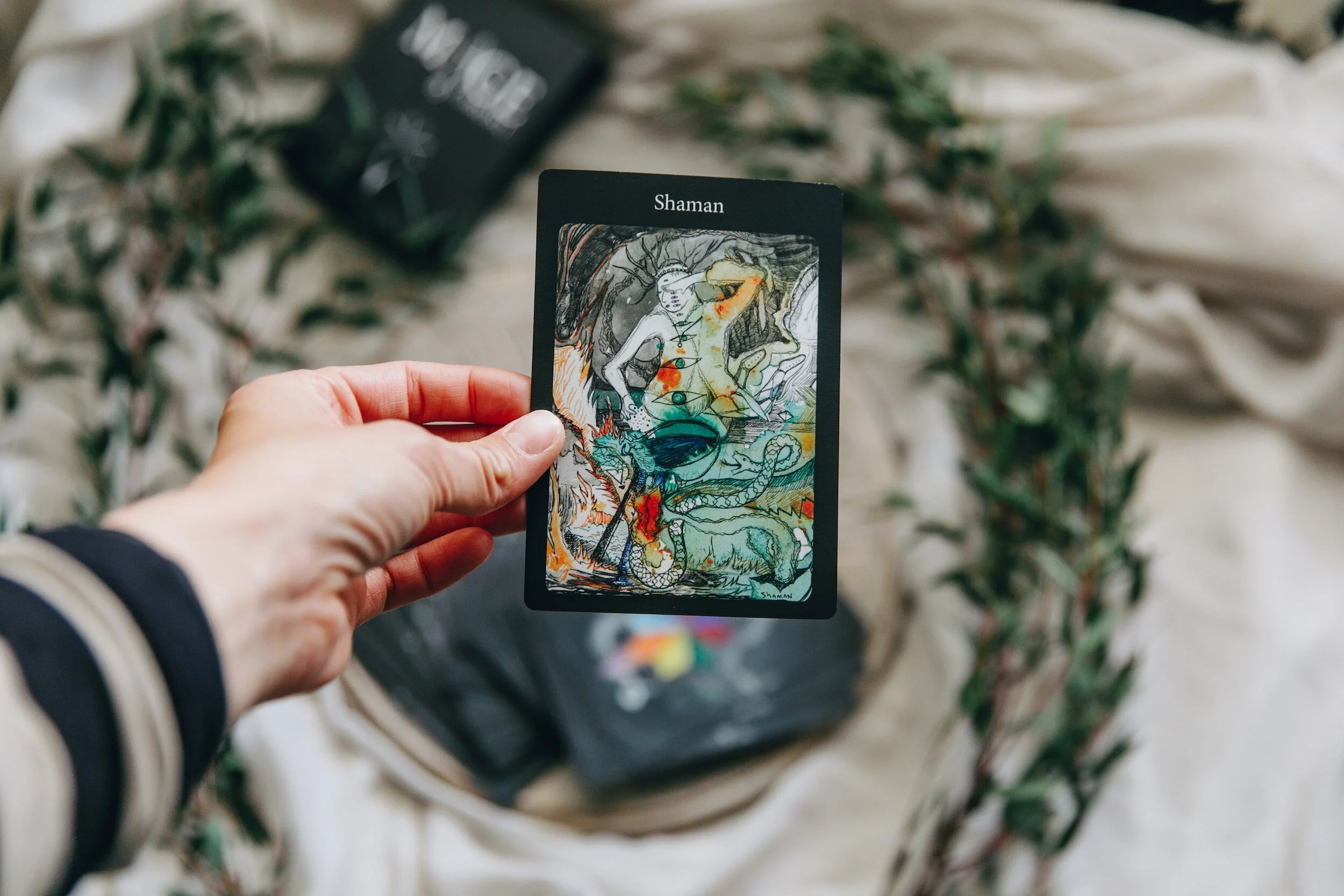 Hand holding a tarot card labeled "Shaman" with abstract artwork of a humanoid figure, mystical symbols, and a dragon on a soft fabric background with greenery.