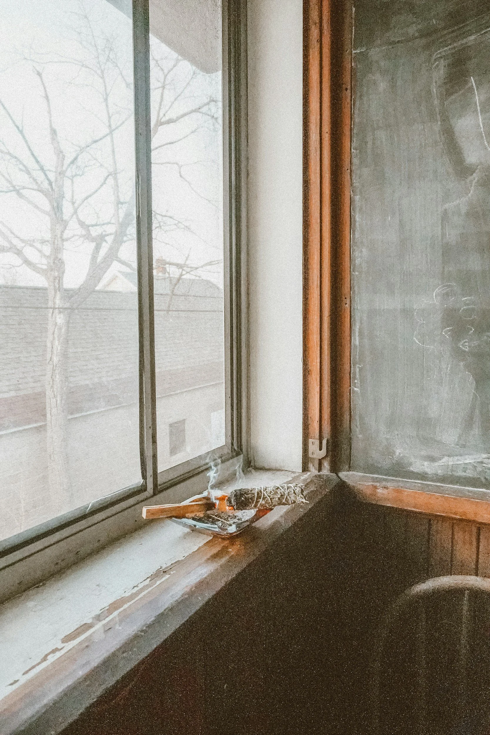 A window with a view of a leafless tree and rooftops, with a small smoldering bundle of herbs or incense smoking on the windowsill next to a chalkboard in a room.