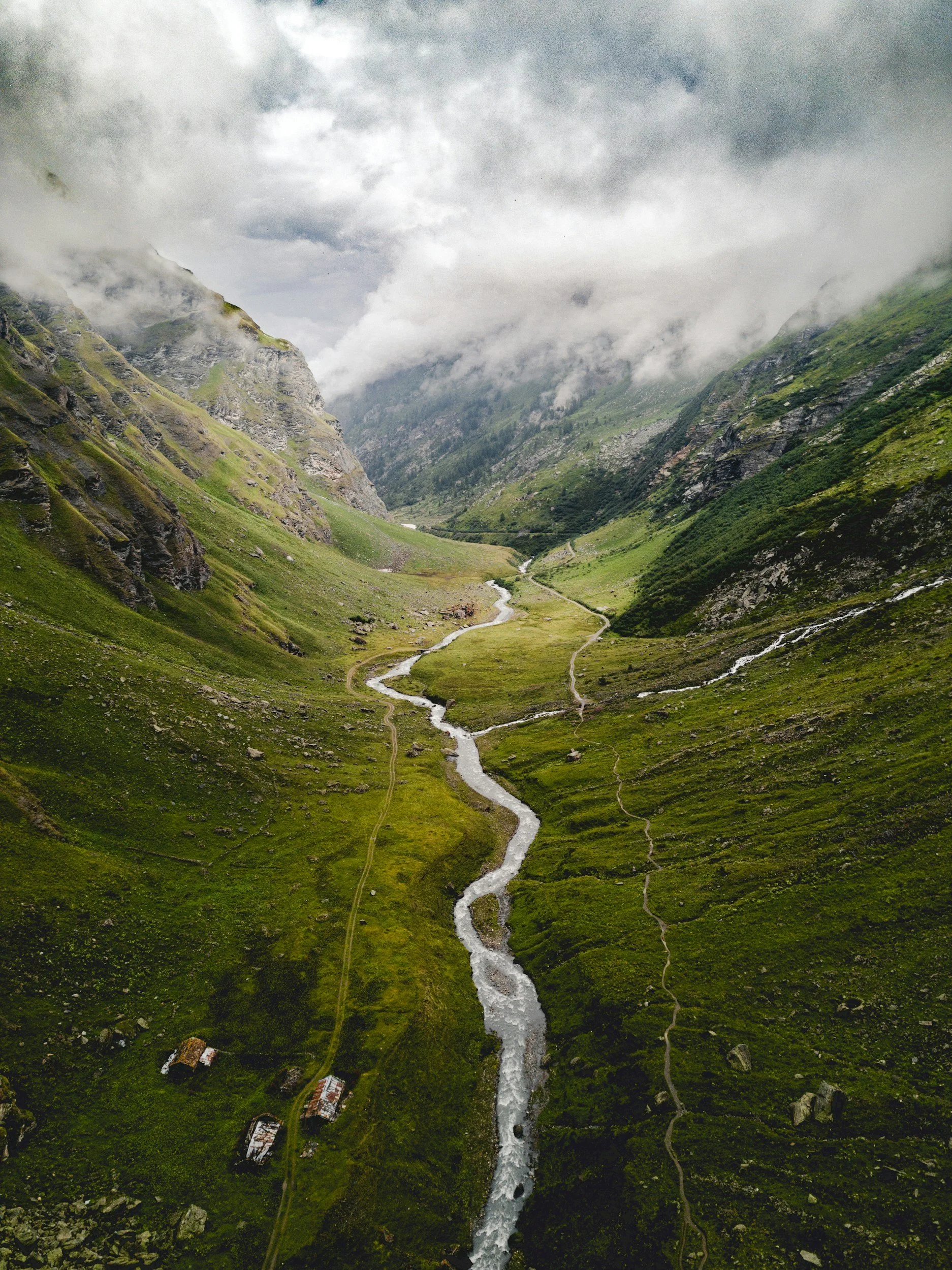 A lush green mountain valley with a winding river and narrow dirt paths, surrounded by misty clouds.