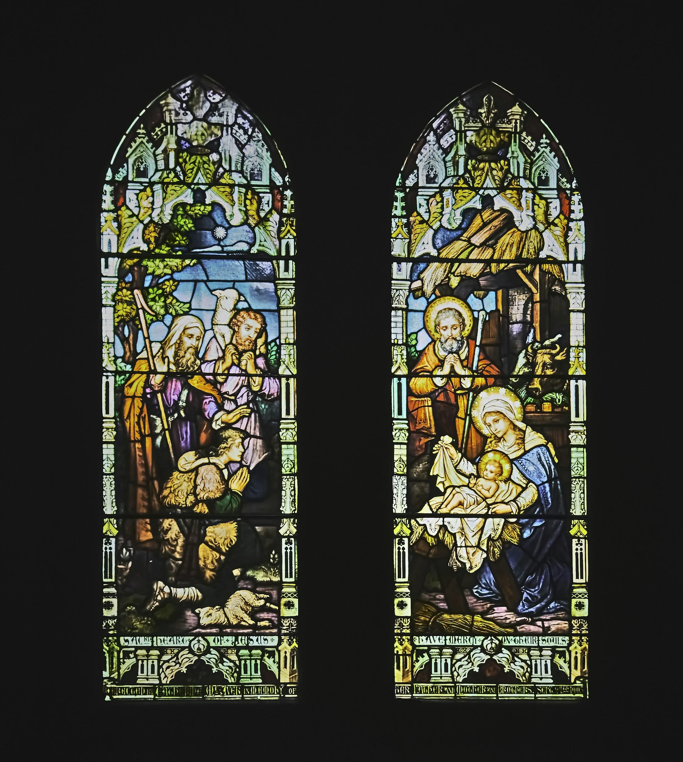 Colorful stained glass windows depicting biblical scenes with saints, a manger, and angels, illuminated against a dark background.