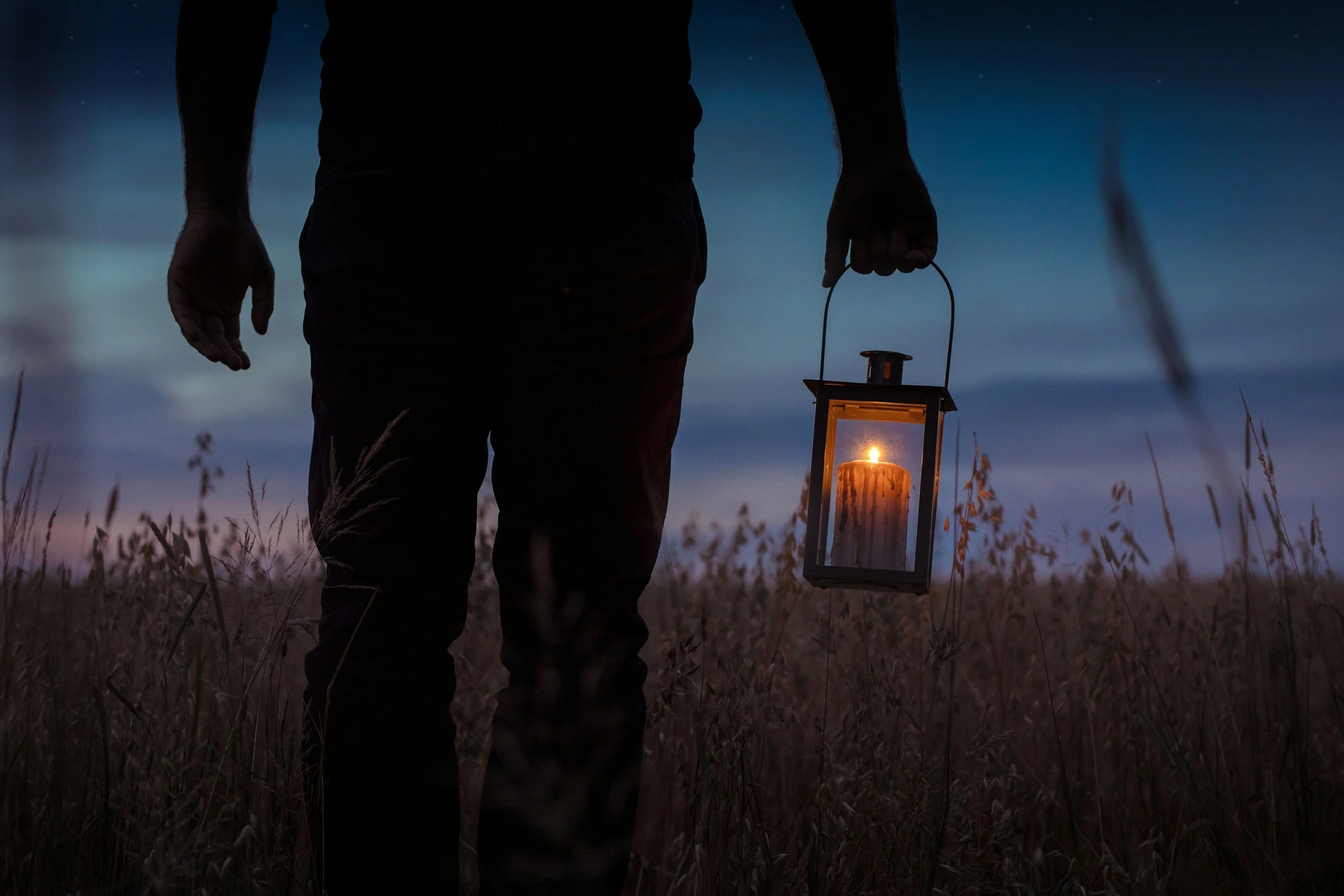 Person walking through a field at dusk holding a lantern with a lit candle inside.