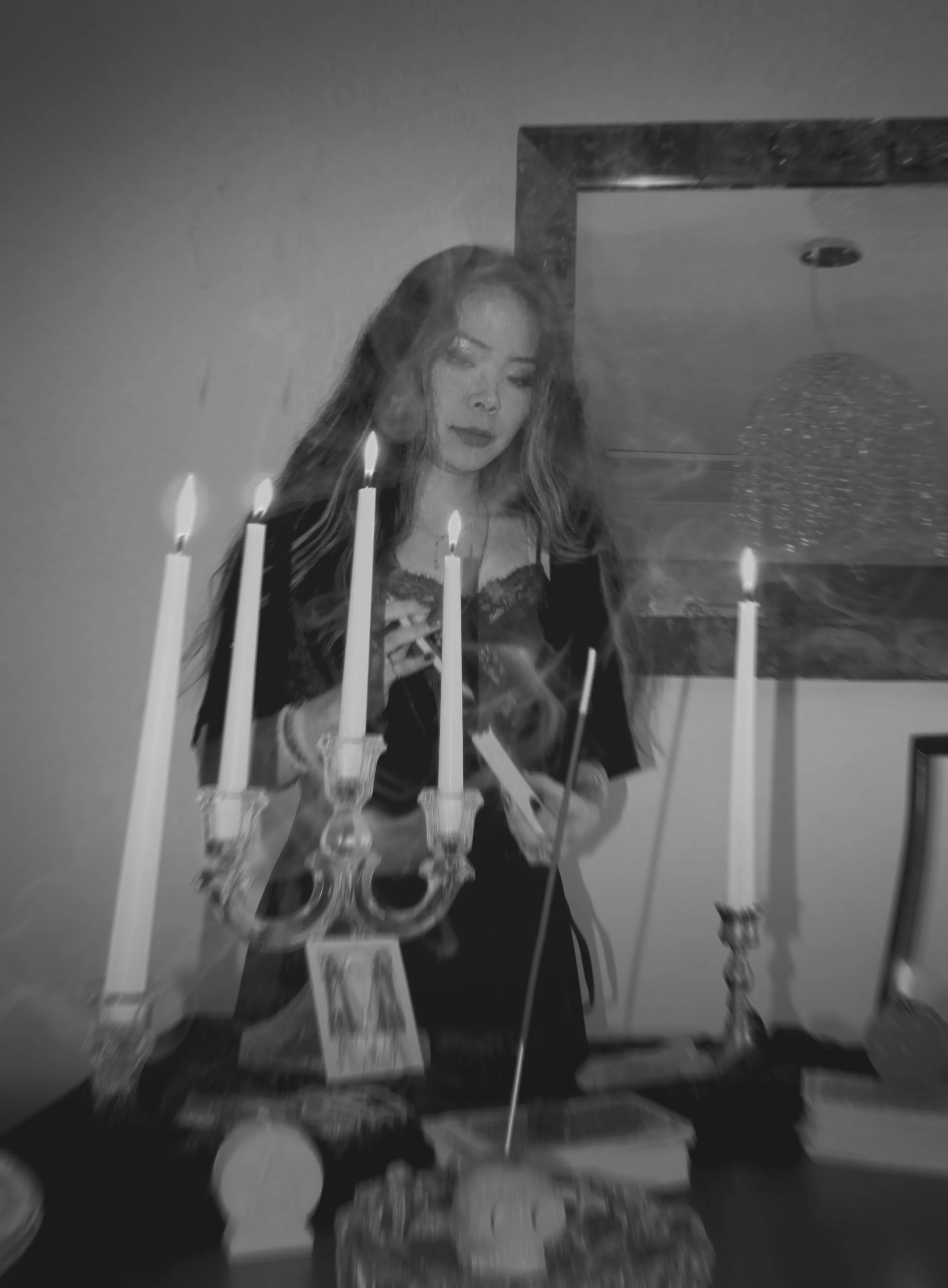 A woman with long hair standing behind a table with lit candles and incense, looking down pensively, with smoke rising around her.