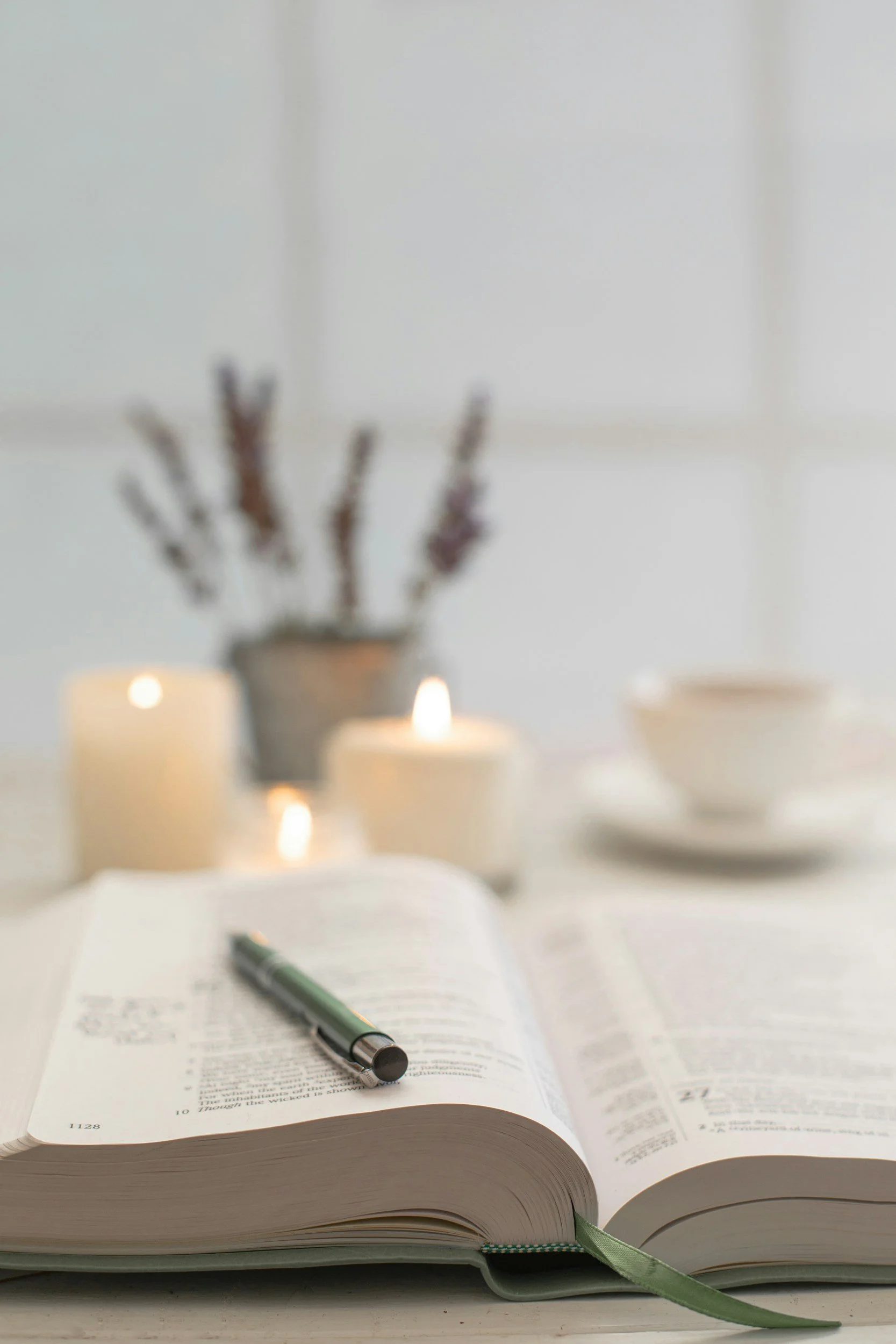 An open book with a pen resting on the pages, with blurred candles and a vase with sticks in the background.