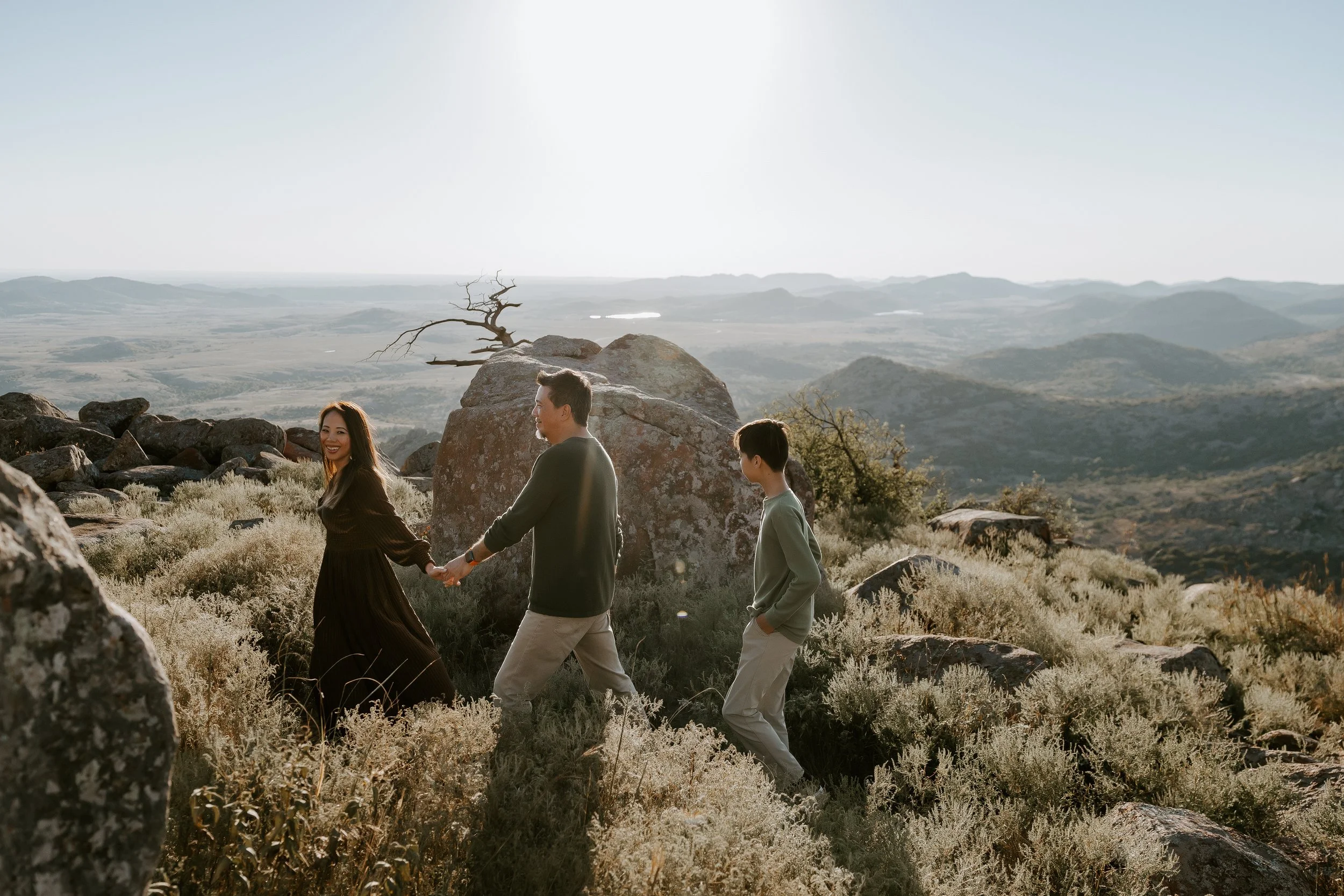 A family of three walking hand in hand in a rocky desert landscape with mountains in the background and bright sunlight overhead.