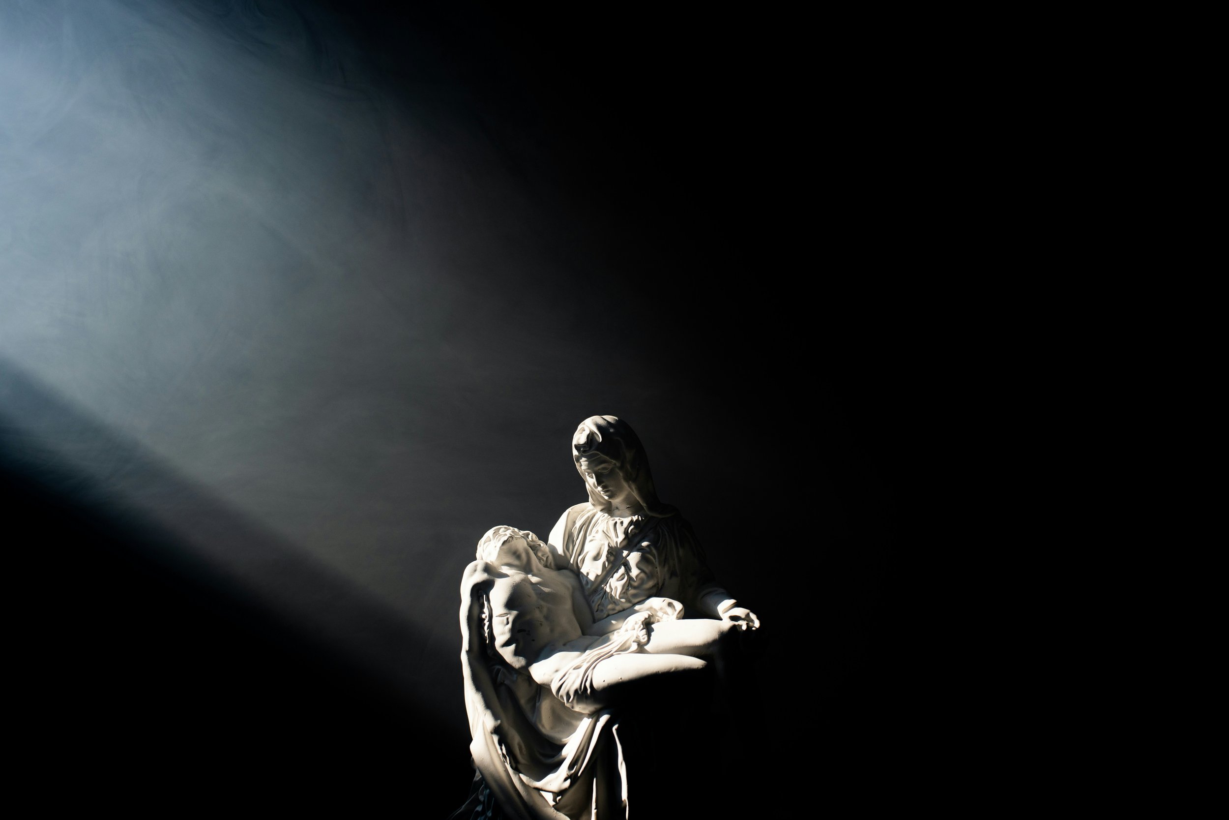 A white marble sculpture of a woman holding a child, illuminated by a dramatic spotlight against a black background.