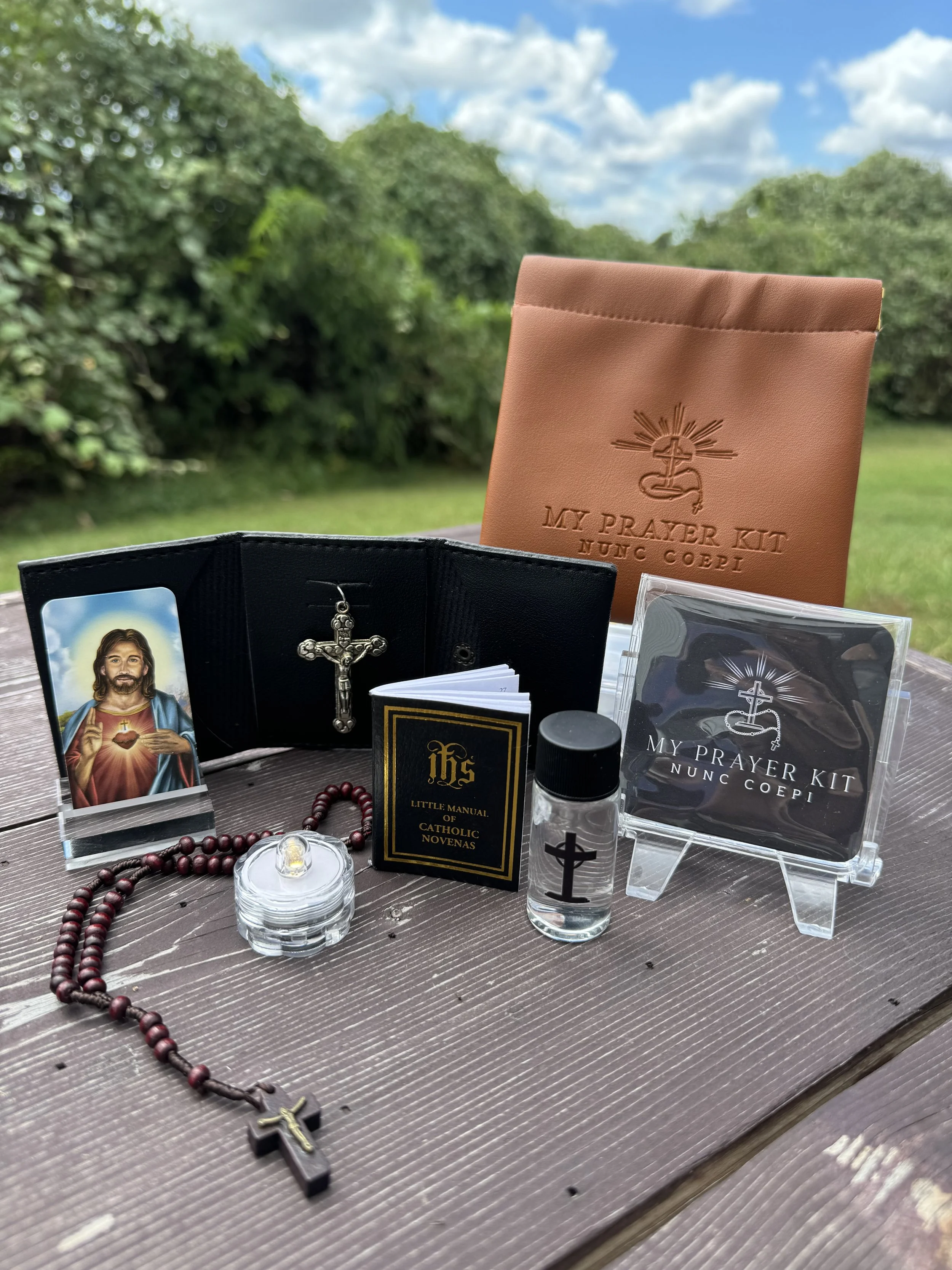 Outdoor scene with religious items including a Jesus image, rosary beads, a crucifix, prayer kits, and religious booklets on a wooden table with a background of green trees and sky.
