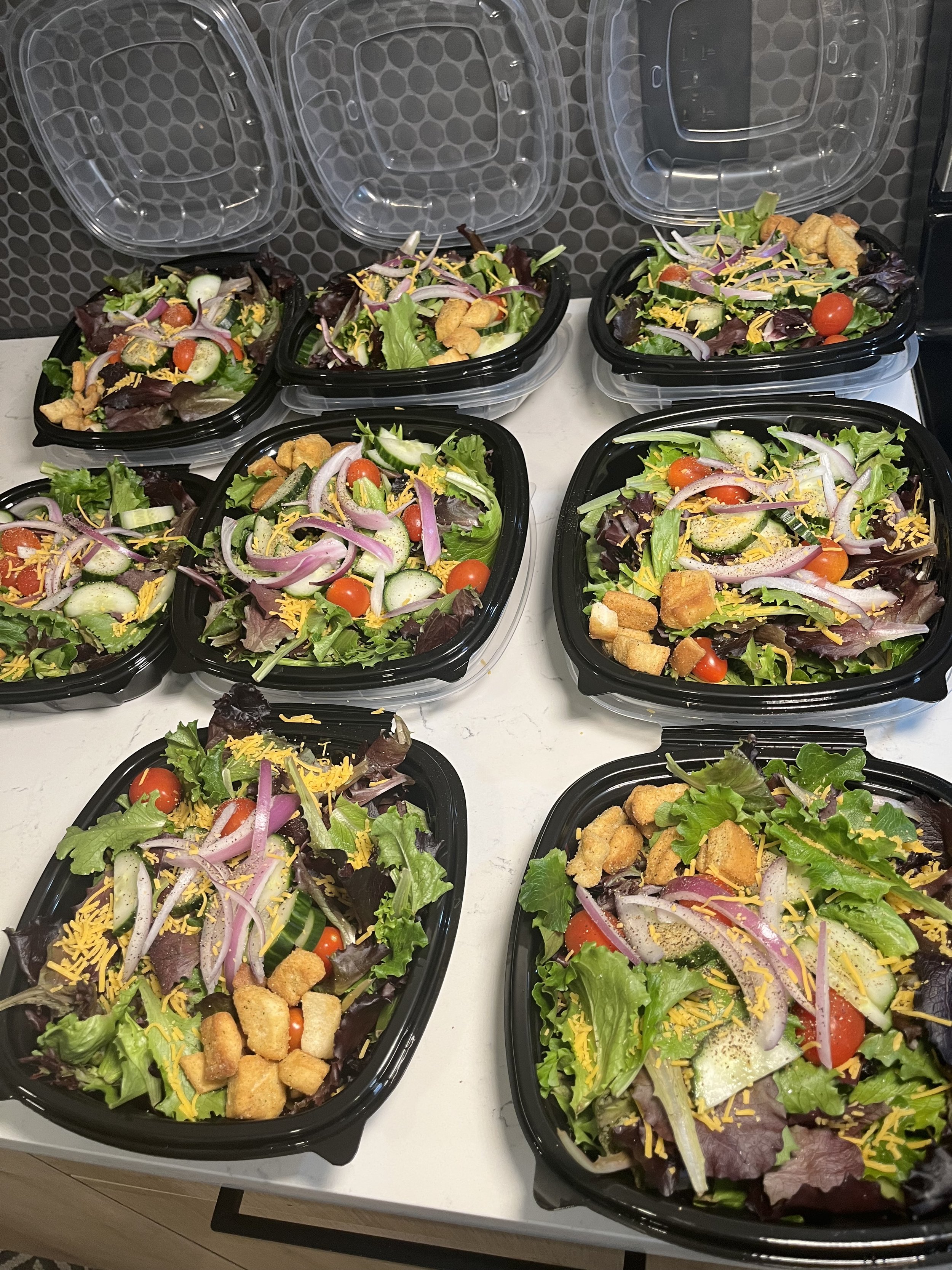 Multiple containers of garden salad with lettuce, cherry tomatoes, sliced cucumbers, shredded cheese, red onion, and croutons.