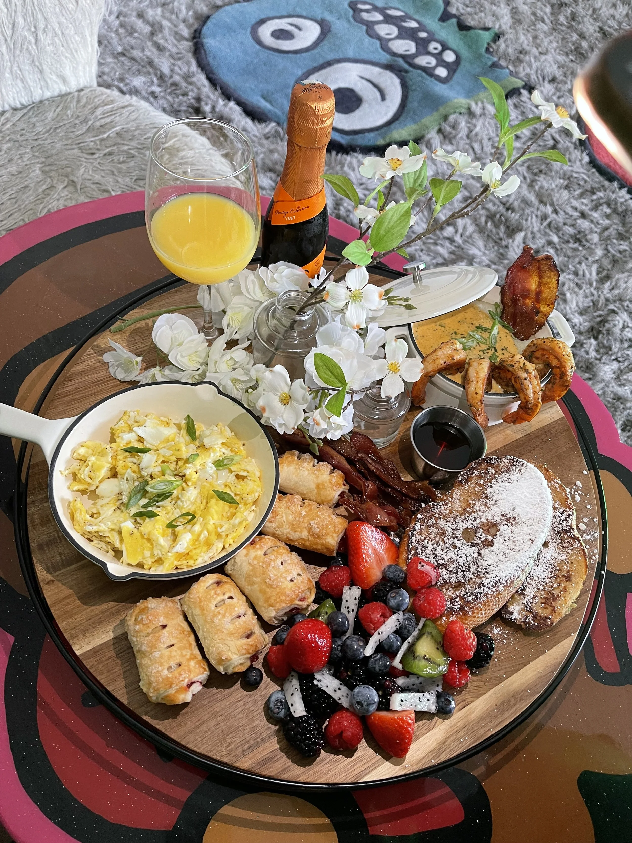 Breakfast platter with scrambled eggs, fruit, pastries, bacon, shrimp, dipping sauce, mimosa, and flowers on a wooden tray.