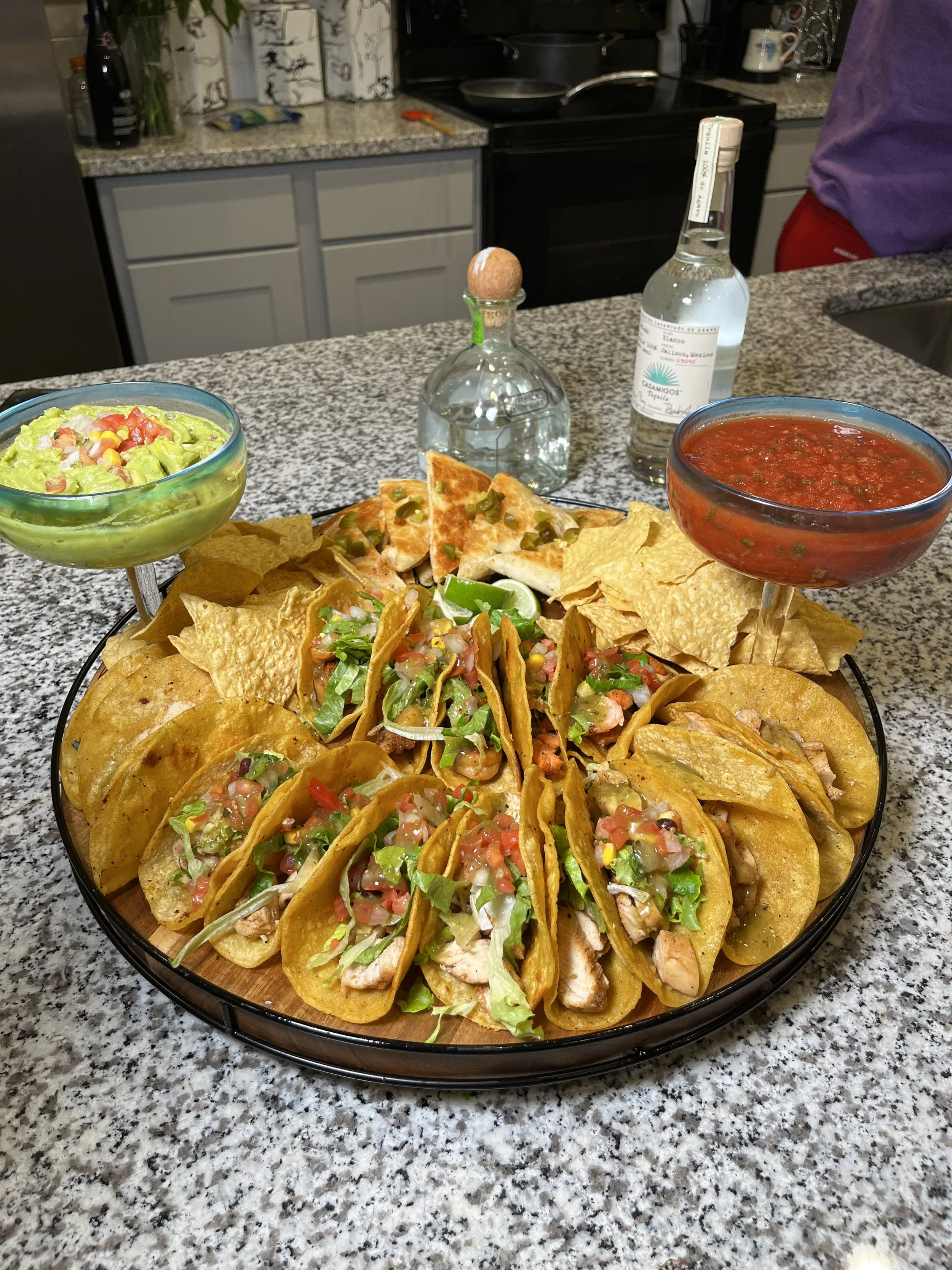 A platter of tacos with lettuce, tomato, and cheese, surrounded by tortilla chips, guacamole, and salsa, on a granite countertop in a kitchen.